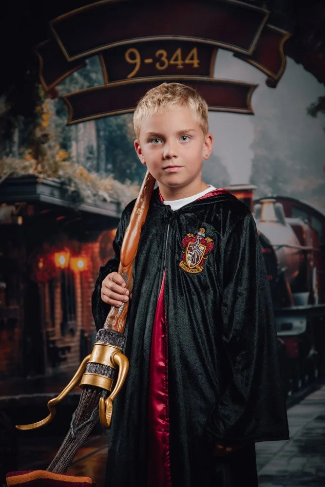Jeune garçon déguisé en héros de Harry Potter, costume noir avec écusson de Gryffondor, tenant une baguette magique, debout devant un décor de train et forêt.