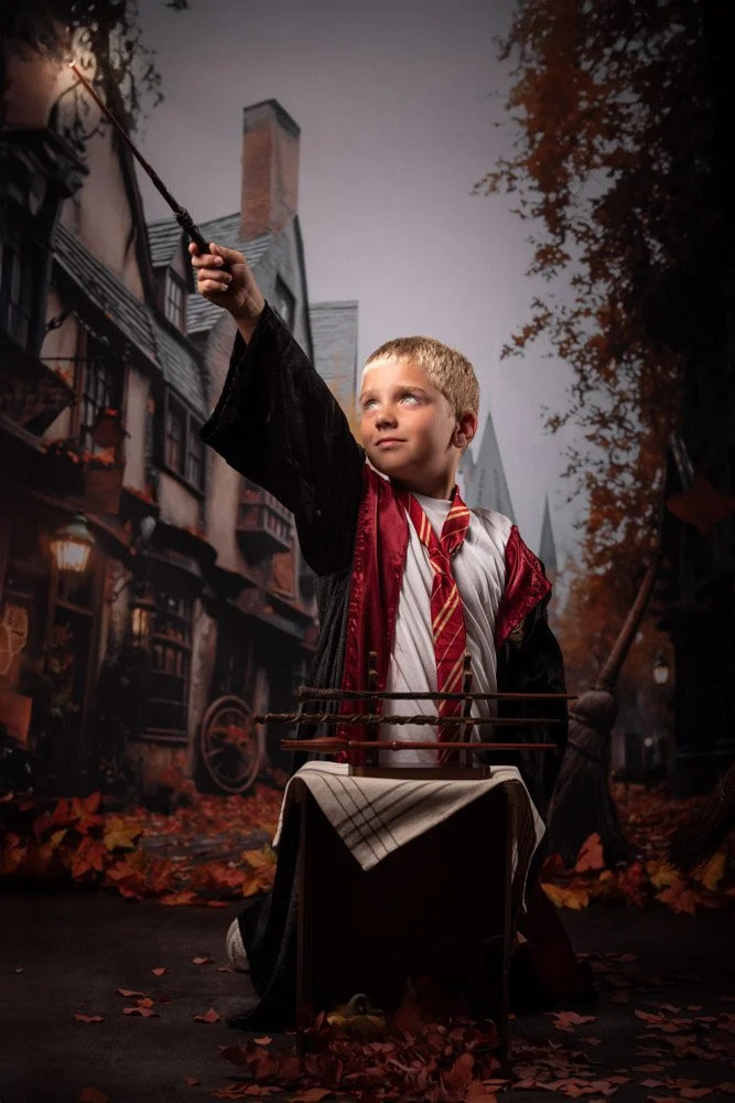 Un garçon habillé en uniforme de Harry Potter, avec une robe et une cravate enroulée, tenant une baguette magique levée, avec un décor évoquant la Borough de Pré-au-Lard de Harry Potter, rempli de feuilles d'automne et de bâtiments anciens.