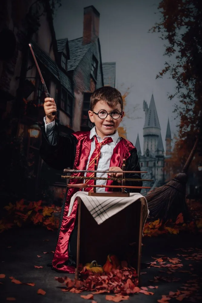 Un jeune garçon déguisé en sorcier avec une baguette magique, portant une robe de sorcier rouge et noir, se tient devant un décor de fantôme avec des bâtiments anciens et une cathédrale en arrière-plan, entouré de feuilles d'automne.