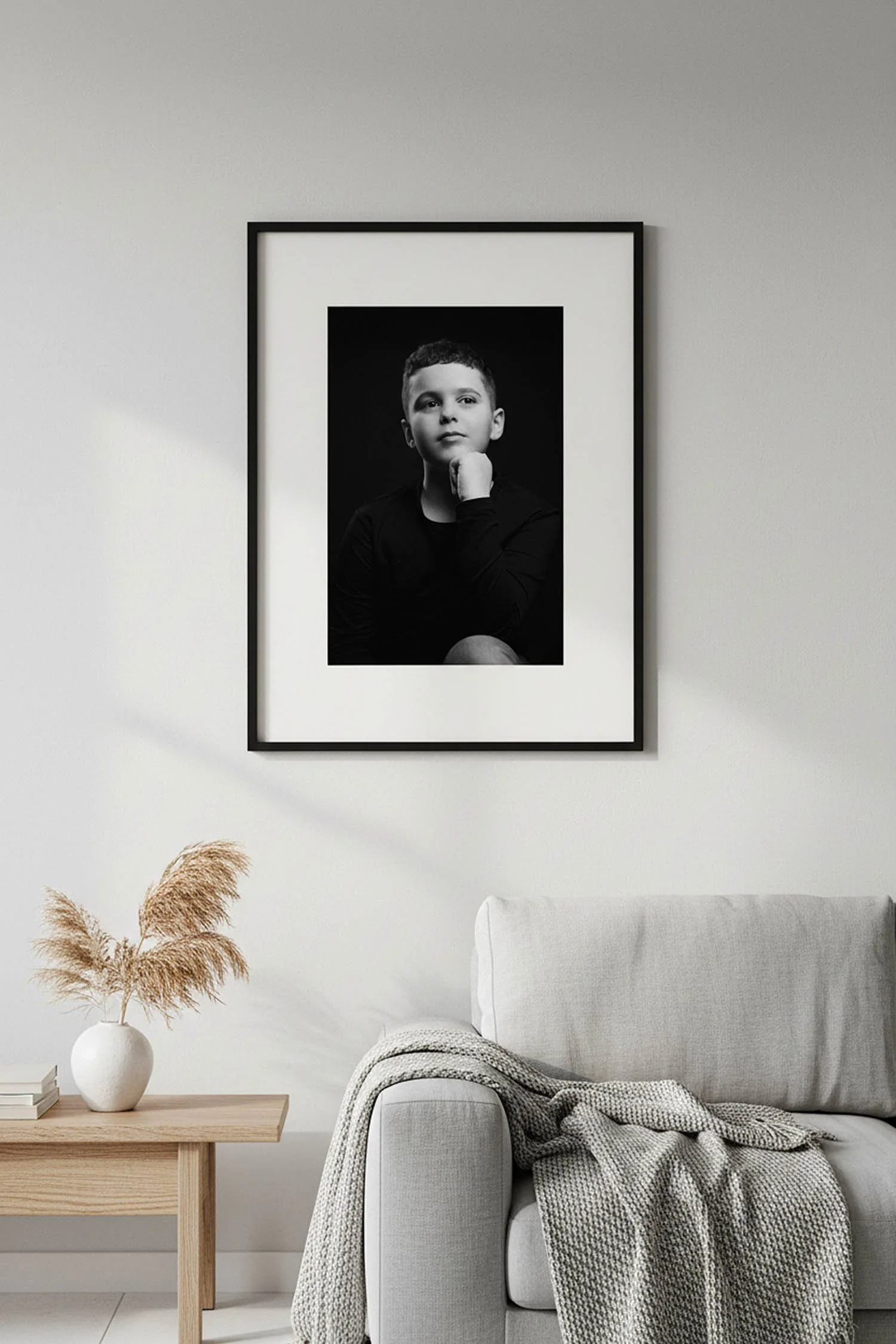 Photo en noir et blanc d'un jeune garçon dans un cadre noir, accroché à un mur blanc au-dessus d'un canapé gris clair, dans un salon minimaliste avec une table en bois clair et un vase avec des herbes pampas