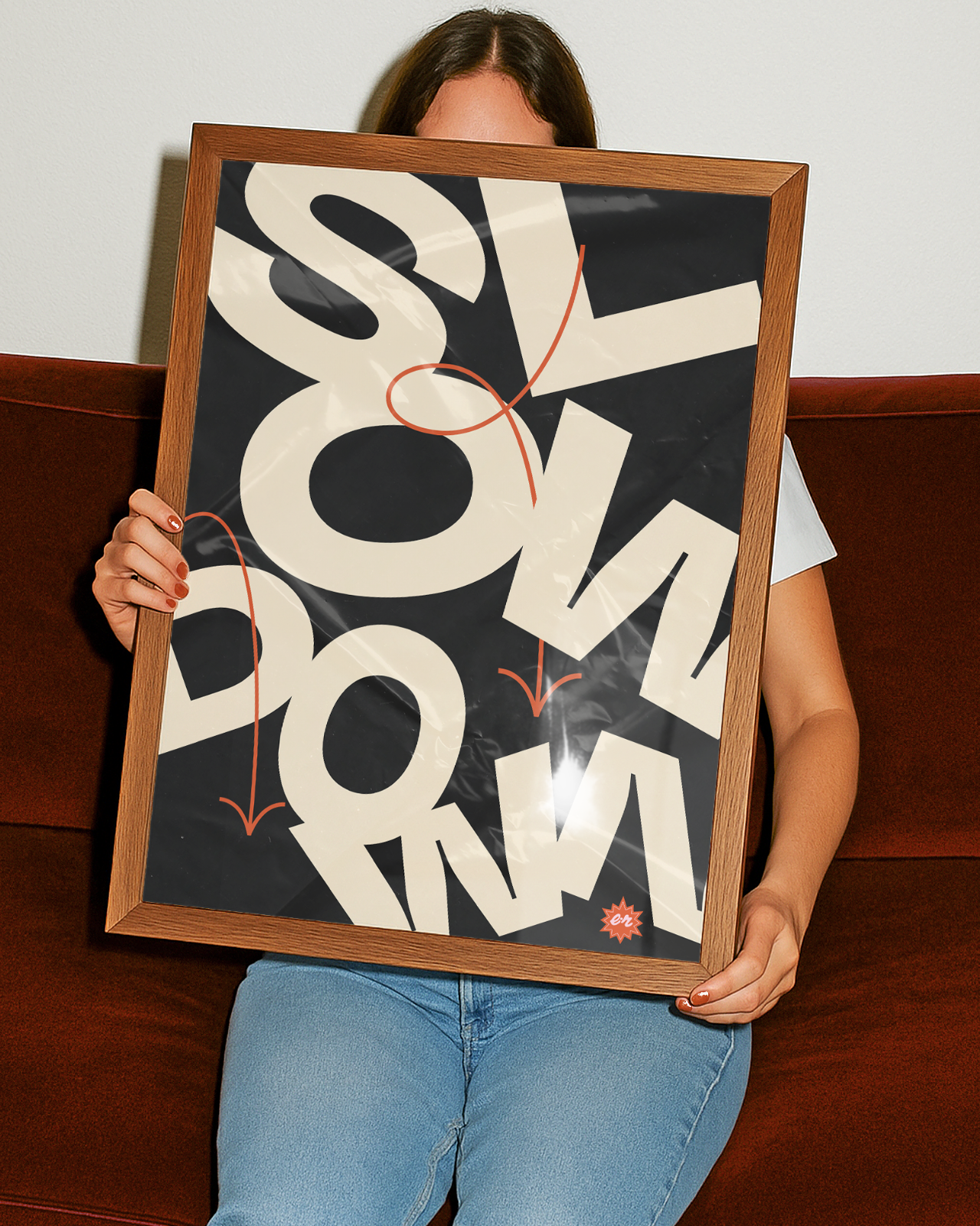 Person sitting on a brown couch holding a large framed poster with bold, overlapping black and cream text and some red graphic elements.