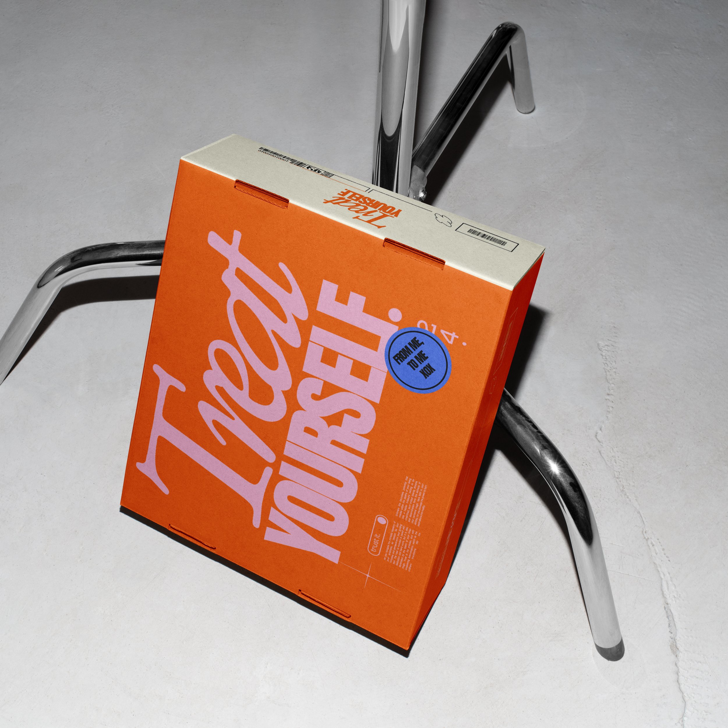 A cardboard box with bright orange and white colors, displaying the text 'Think Yourself' and 'From Me to You Now' on a metal chair with a curved chrome frame, placed on a light concrete floor.