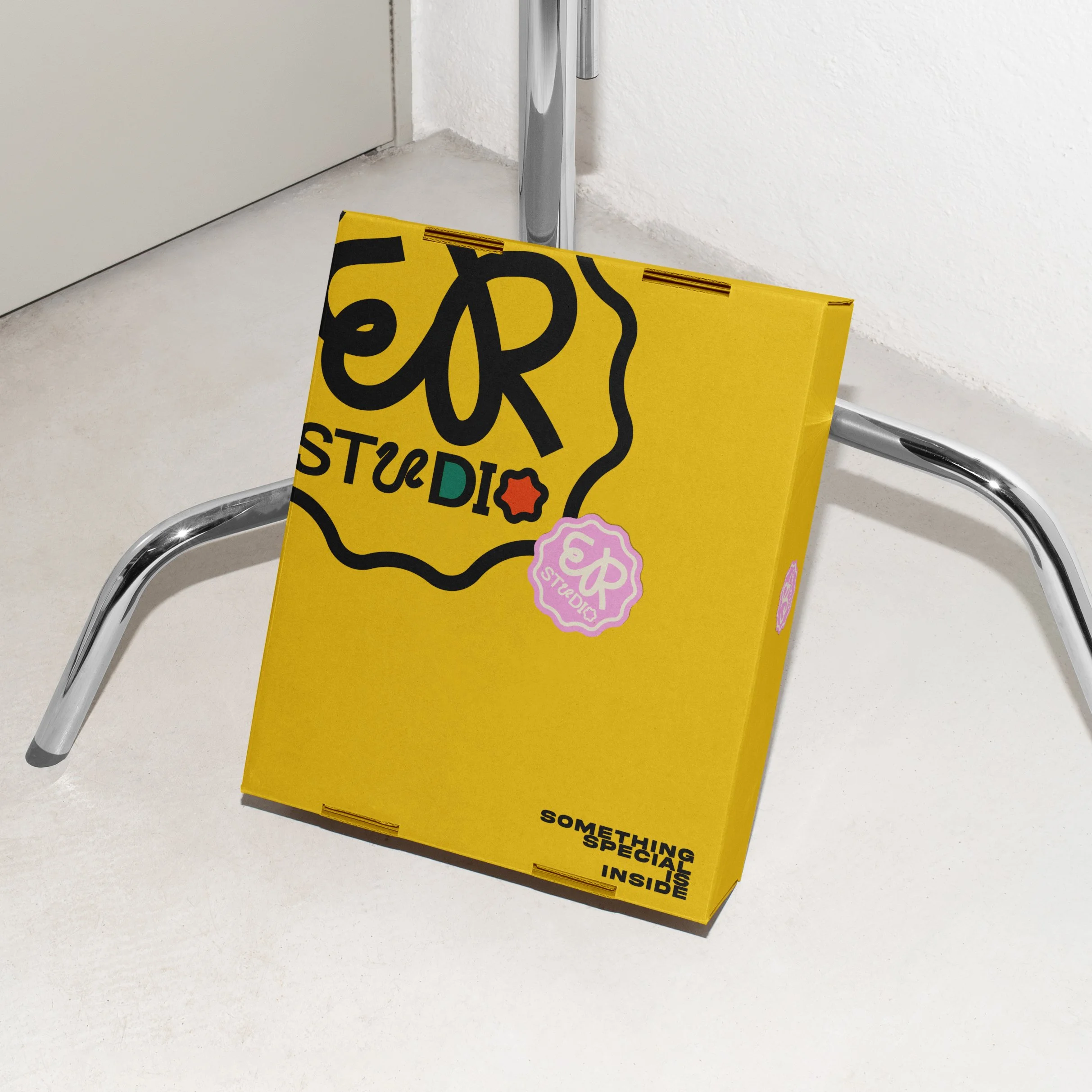 Yellow cardboard box with colorful text and graphics leaning against a metal hand truck on a white floor.