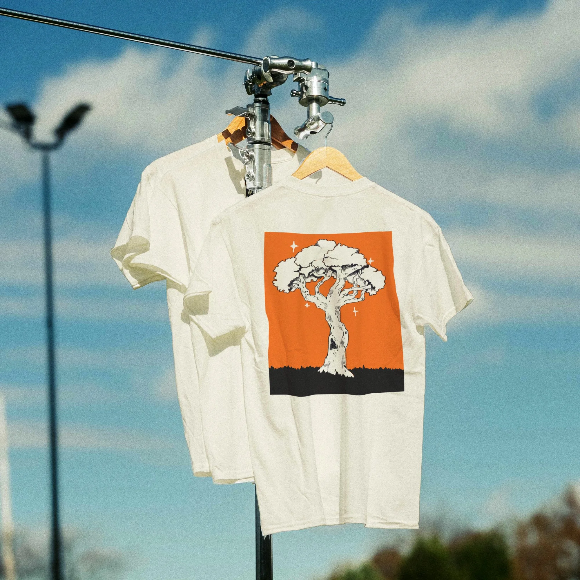Two white T-shirts hanging on a metal rack outdoors, one with a tree illustration on an orange background, under a blue sky.