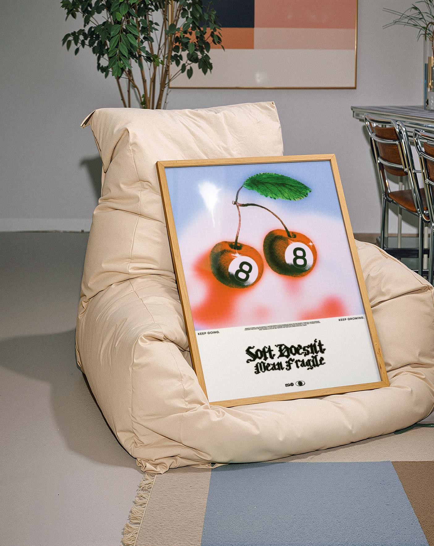 A framed poster of two cherries with the number 8 on each, leaning against a beige bean bag chair in a modern room with a potted plant and a dining set in the background.