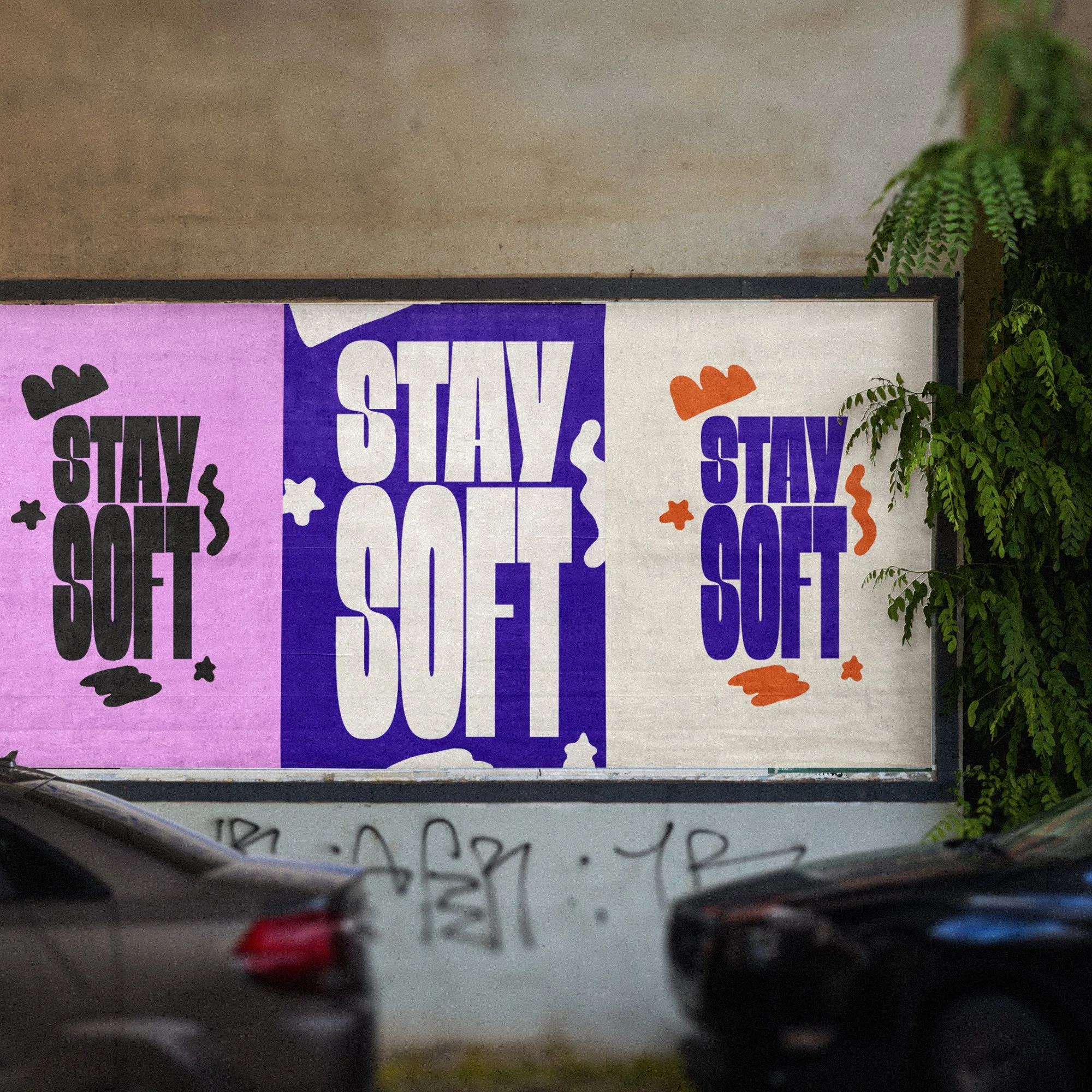 A large outdoor billboard with three panels displaying the phrase 'STAY SOFT' in bold capital letters, with decorative elements, colorful backgrounds, and green foliage on the right side.