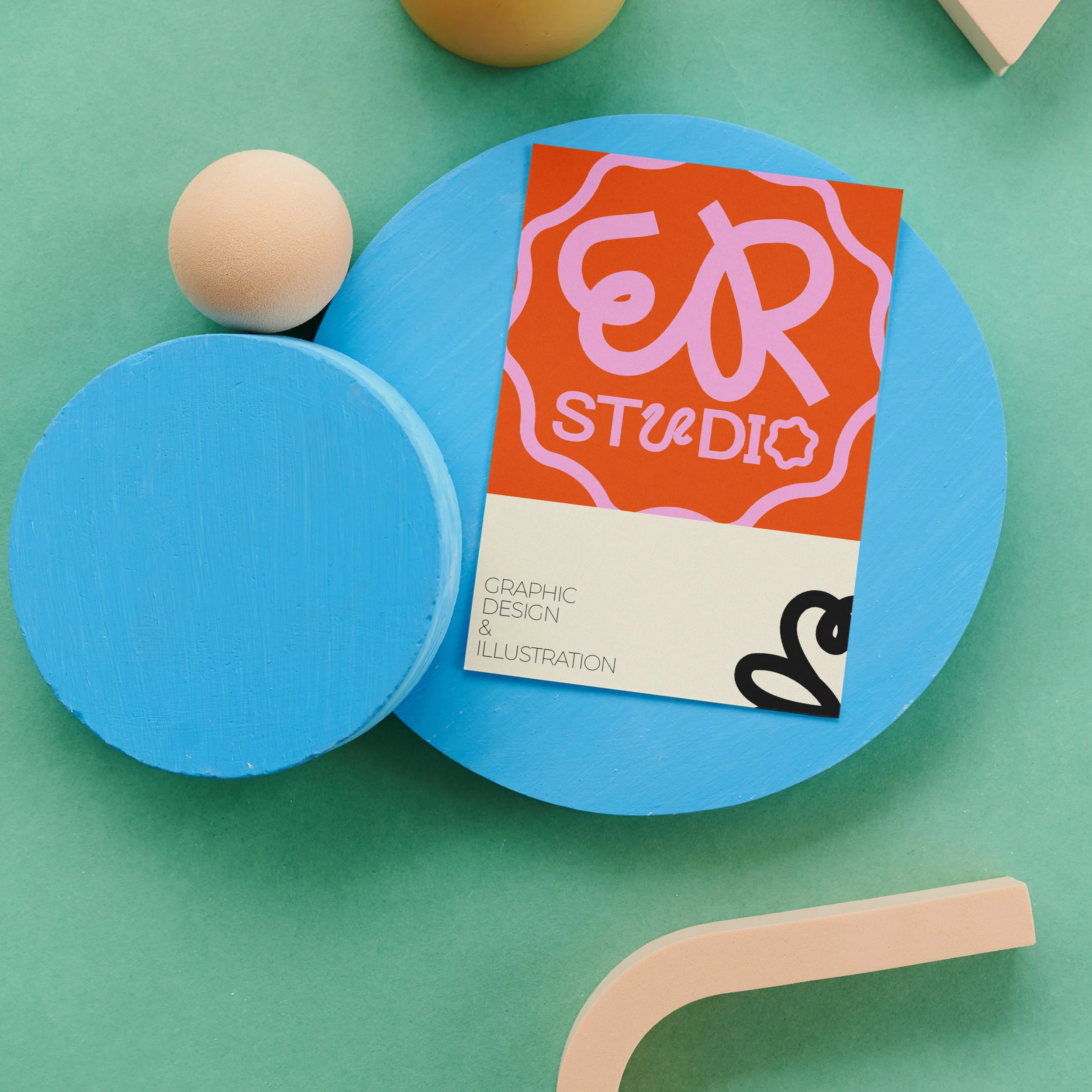 Colorful graphic design and illustration studio business card placed on two blue circular platforms, with a foam ball and a light pink object nearby, on a pastel green background.
