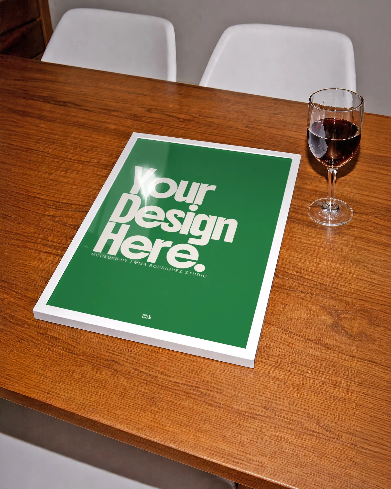 A green mockup poster on a wooden table with the text 'Your Design Here. Mockups by Emma Rodriguez Studio' and a glass of red wine beside it.