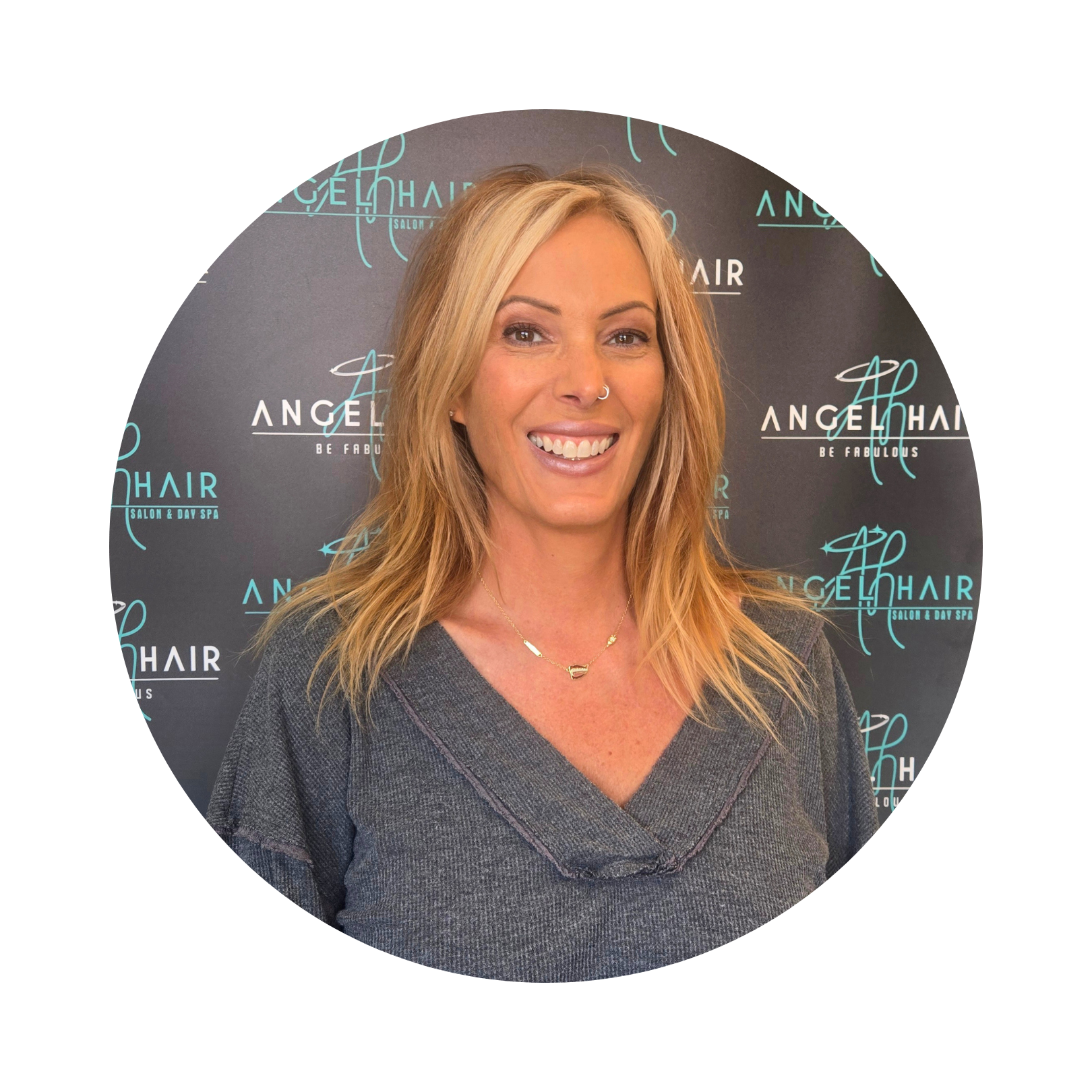 A woman with blonde hair and a nose ring smiling at the camera, standing in front of a backdrop with the logo of Angel Hair Salon and Day Spa.