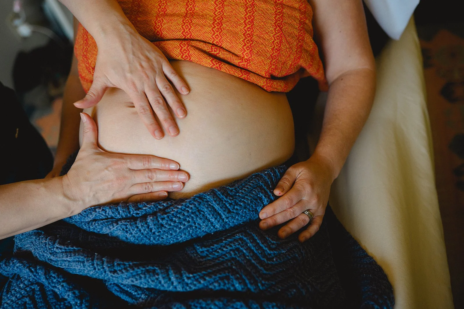 A person receiving a massage or physical therapy, with two hands on their bare abdomen, one on top and one on the side. The person is wearing a colorful orange top, and a blue knitted garment is around their waist.