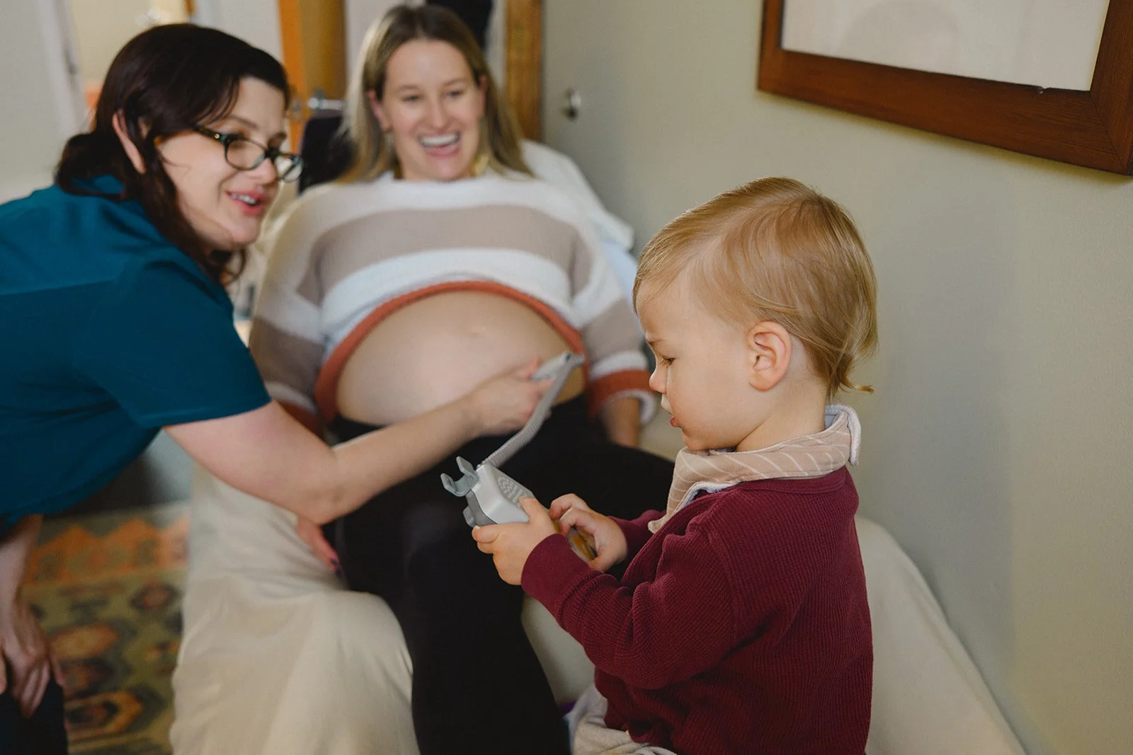 A pregnant woman in a hospital bed smiling, with a young boy holding a toy and looking at her abdomen, and a woman with glasses leaning over smiling.