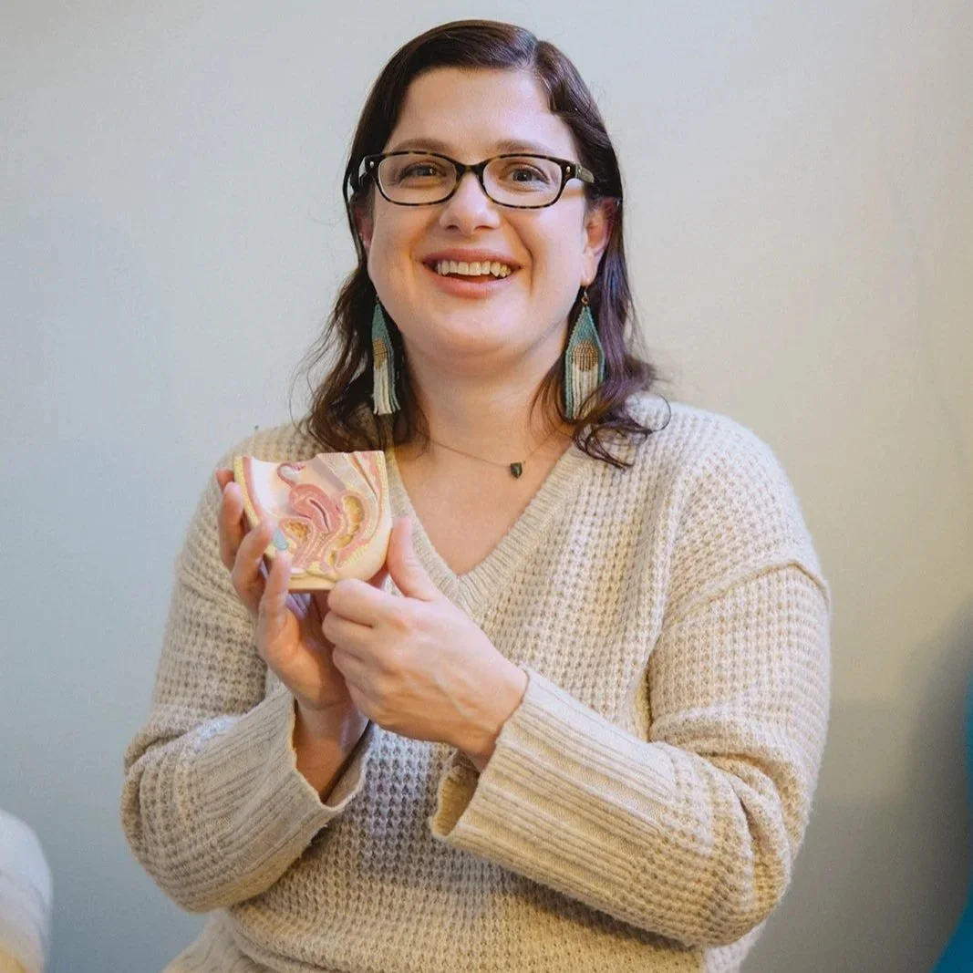 A woman with glasses and earrings holding a soap with colorful swirls, smiling in front of a plain wall.