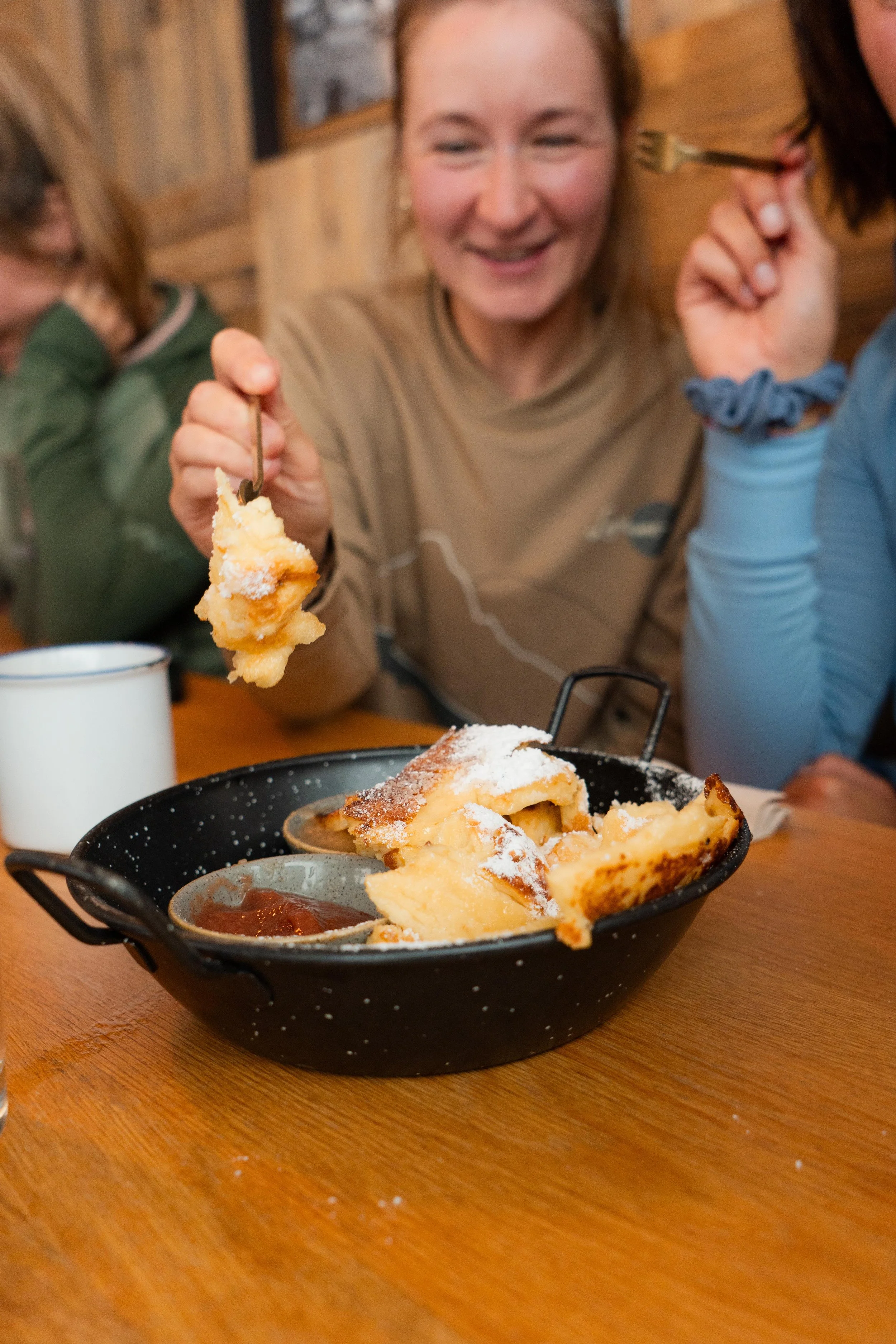 Eine lachende Frau hält ein Stück Auflauf oder Bienenstich mit einem Löffel, vor ihr auf dem Tisch ein großer, halb gegessener Kuchen mit Puderzucker und Marmelade in einer schwarzen Schüssel.
