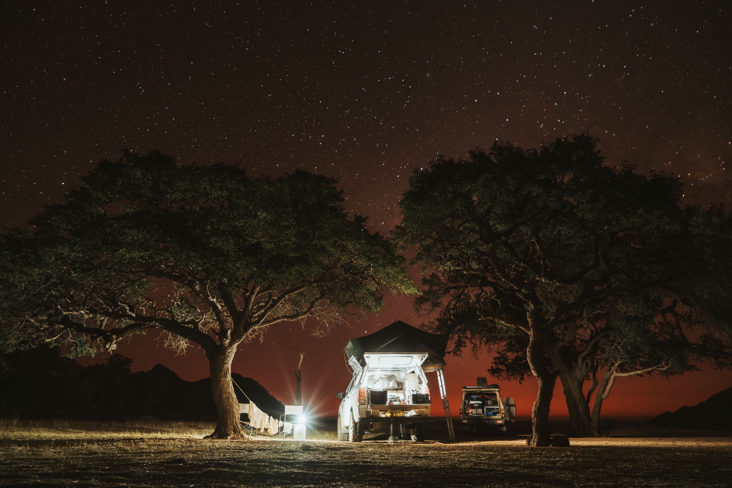 Campingplatz unter einem Sternenhimmel bei Dämmerung, mit zwei großen Bäumen, einem Autokanu mit offener Heckklappe und einem zweiten Fahrzeug, auf dem ein Dachzelt steht, im Wüsten- oder Gebirgsraum.