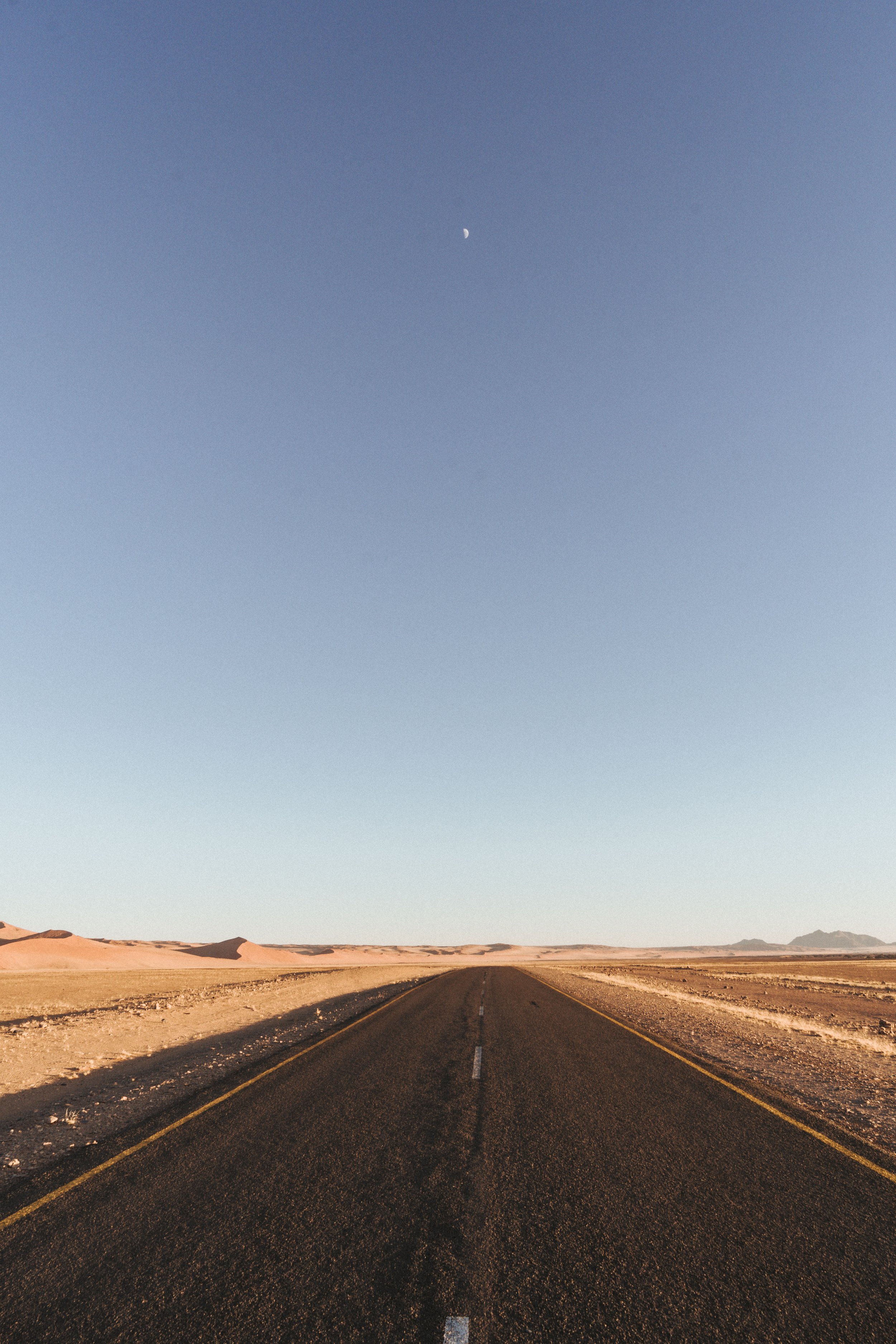 Eine einsame Straße in einer Wüste mit Berge im Hintergrund und einem blauen Himmel mit sichtbarem Mond.