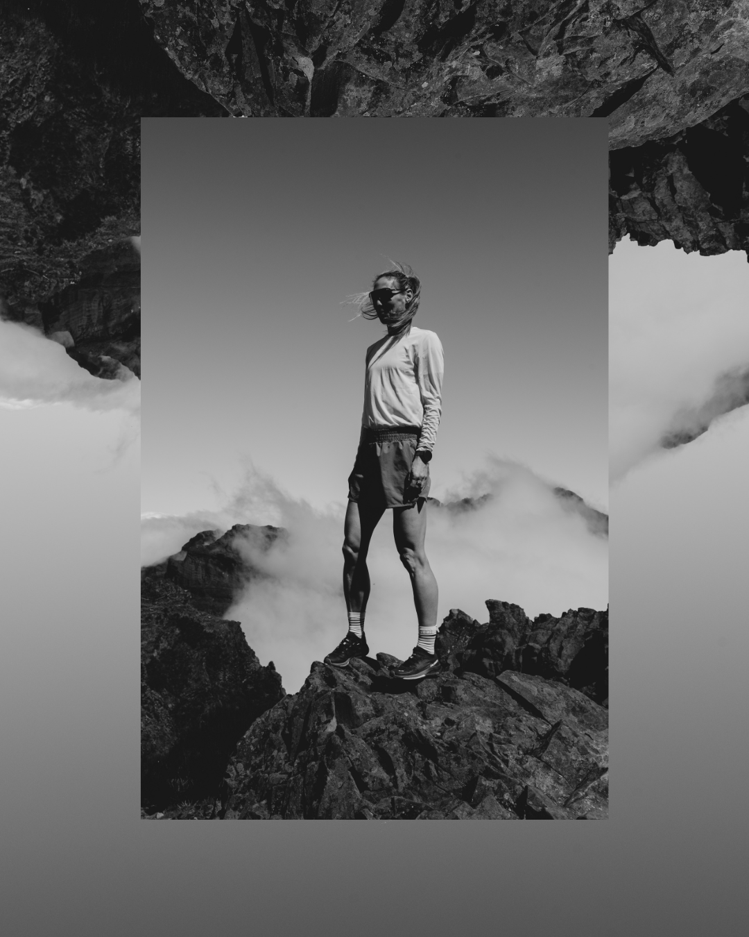 Schwarz-weiß Fotografie einer jungen Frau, die auf einem Felsen in den Bergen steht, mit Wolken und Himmel im Hintergrund.