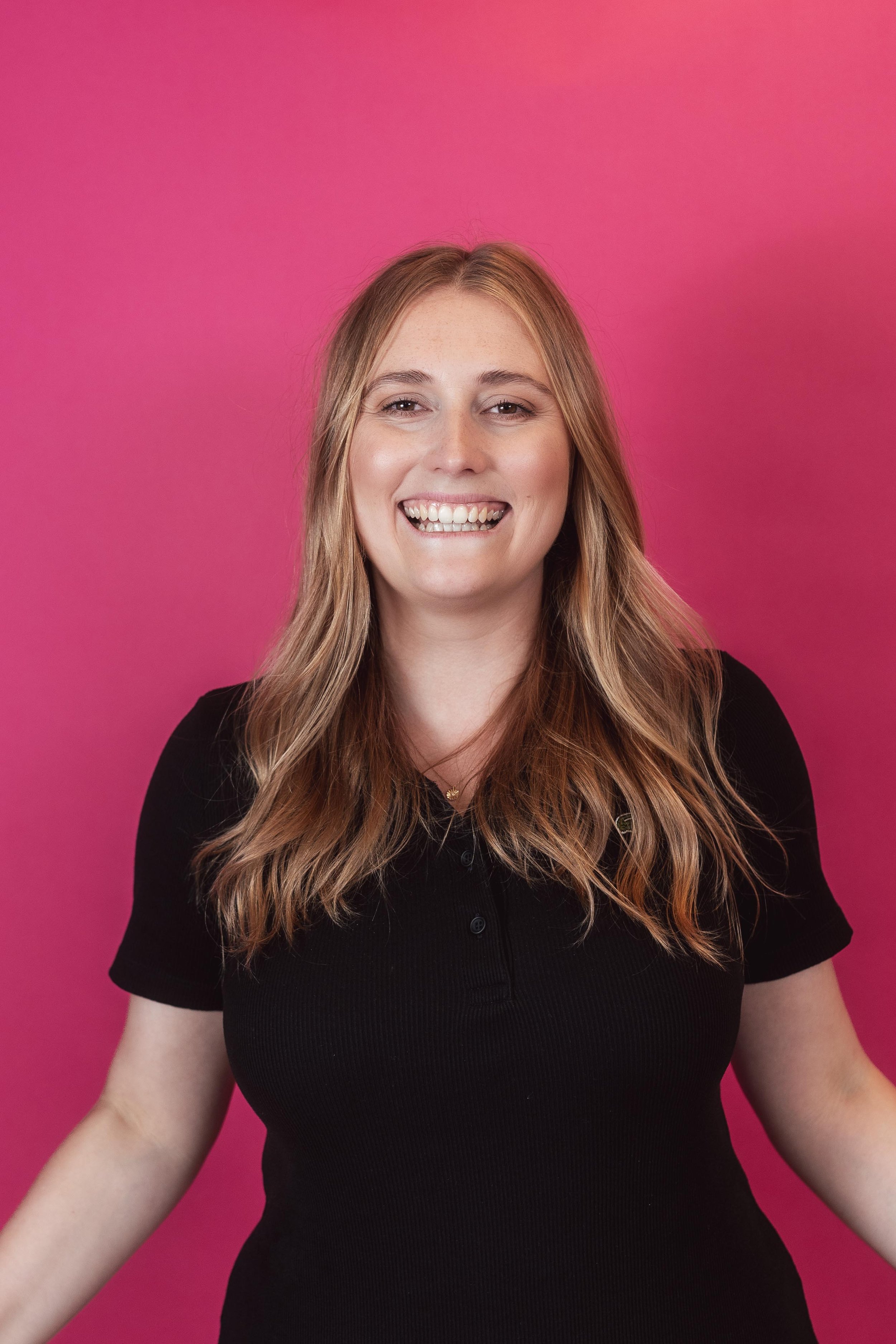 Junge Frau mit langen, gewellten Haaren, die in die Kamera lächelt, vor pinkem Hintergrund, trägt schwarzes T-Shirt.
