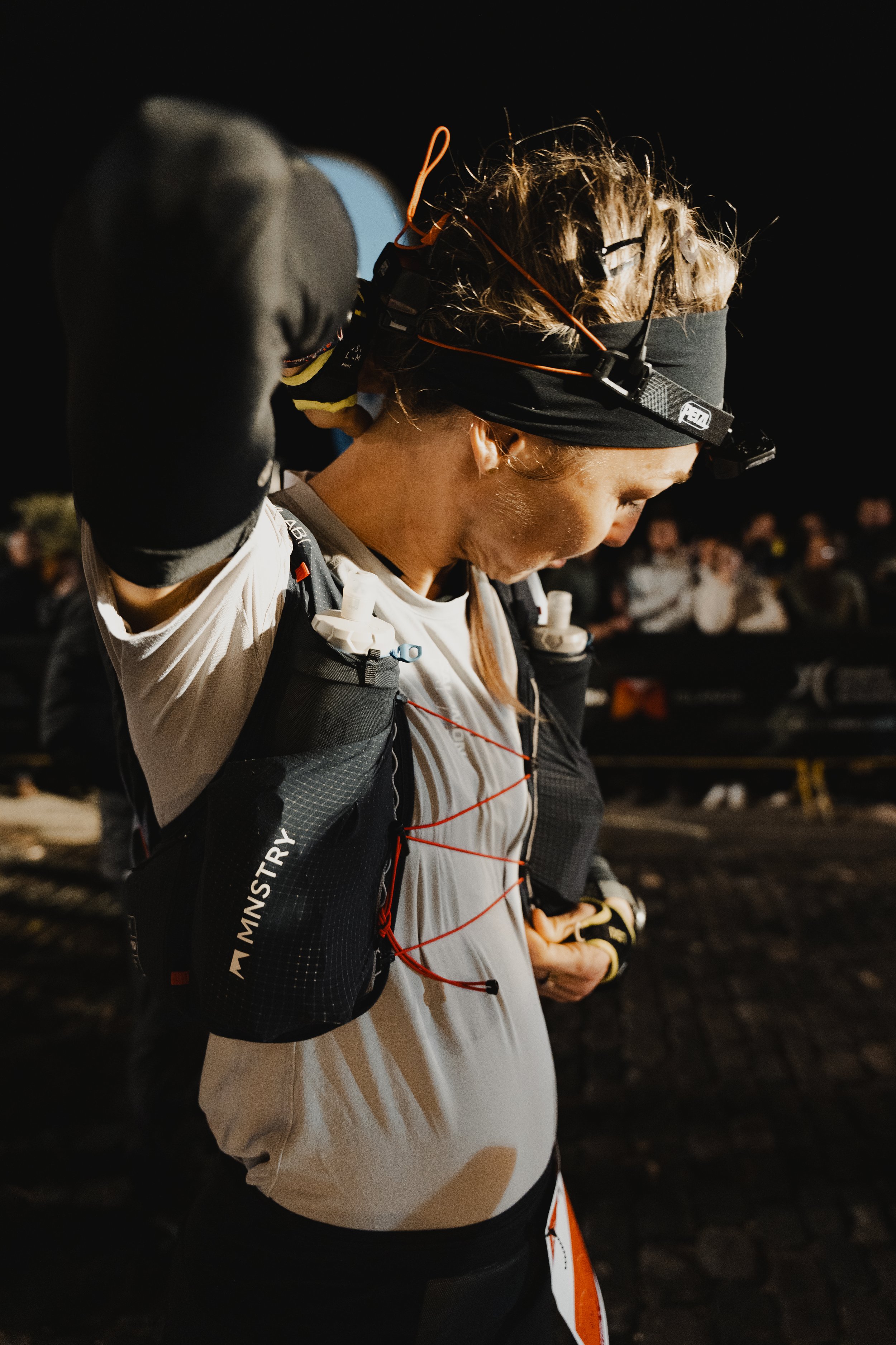 Frau bei Nacht beim Trailrunning, trägt Kopflampe und Sporttasche, in einer Menschenmenge.