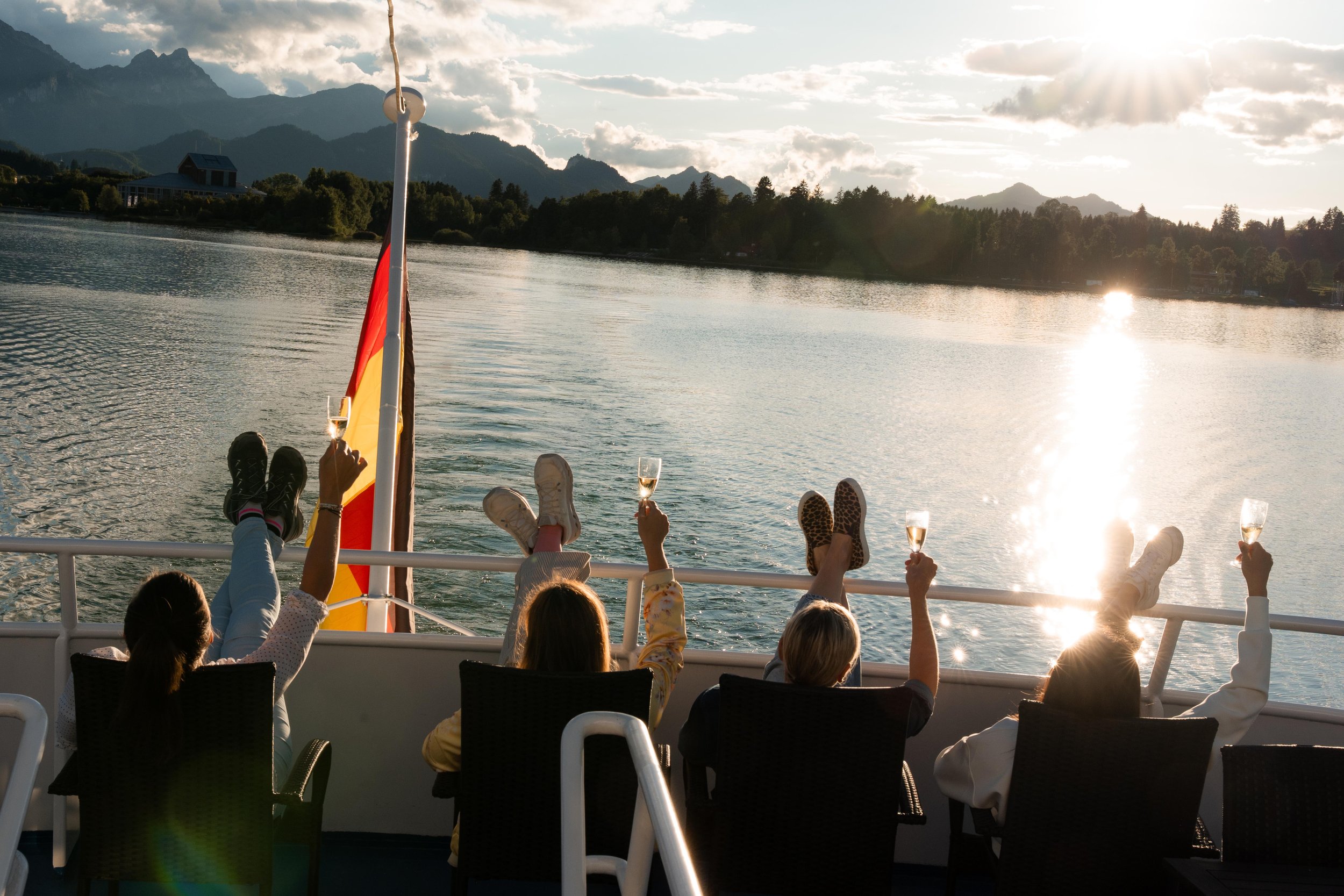 Menschen auf einem Boot, liegend mit erhobenen Beinen, die Wein trinken, bei Sonnenuntergang auf einem See mit Bergen im Hintergrund.