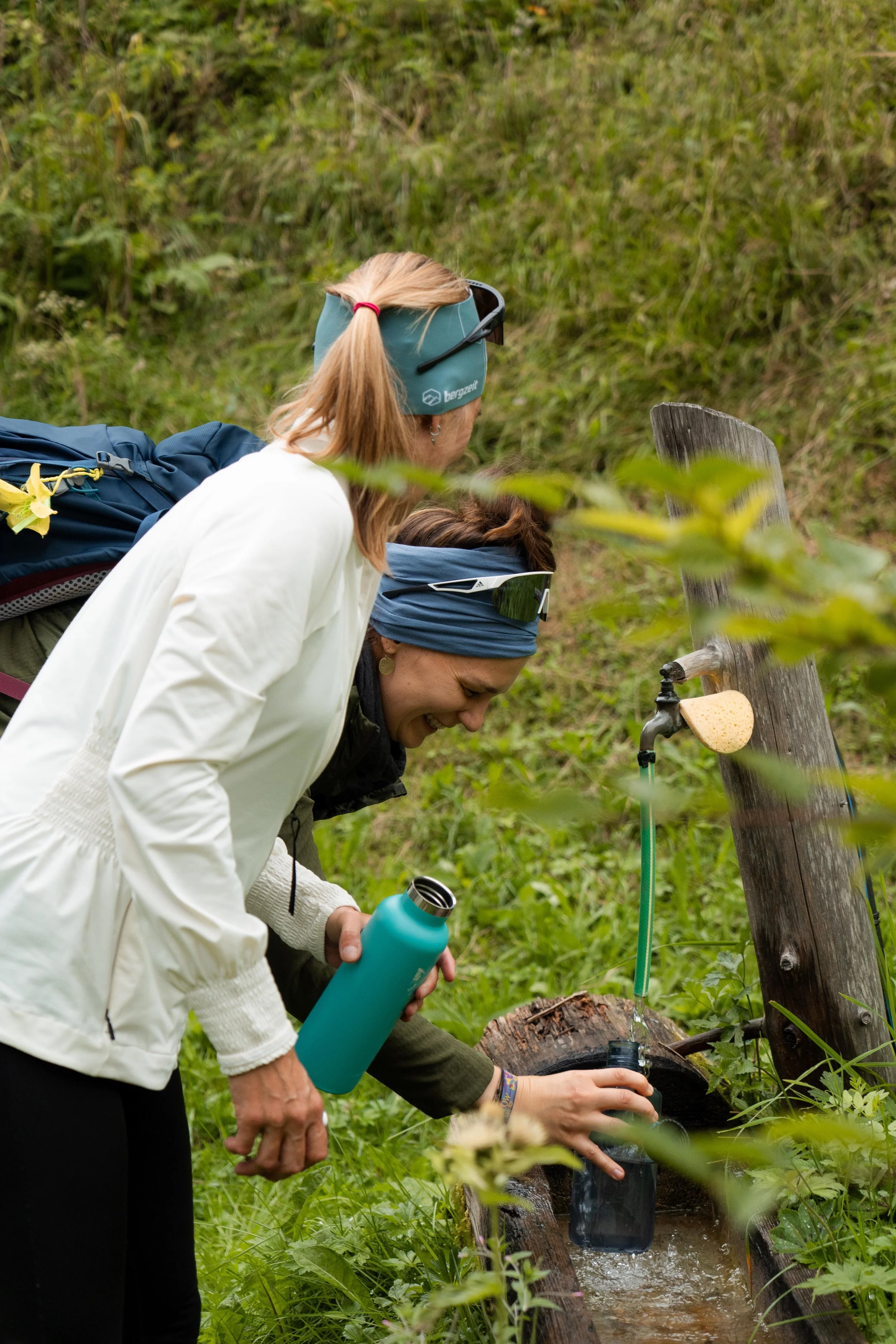 Zwei lächelnde Frauen füllen eine Trinkflasche an einer Wasserquelle in der Natur auf, umgeben von grünem Laub. Eine der Frauen trägt eine blaue Stirnband und die andere einen blauen Kopfschutz mit Sonnenbrille.