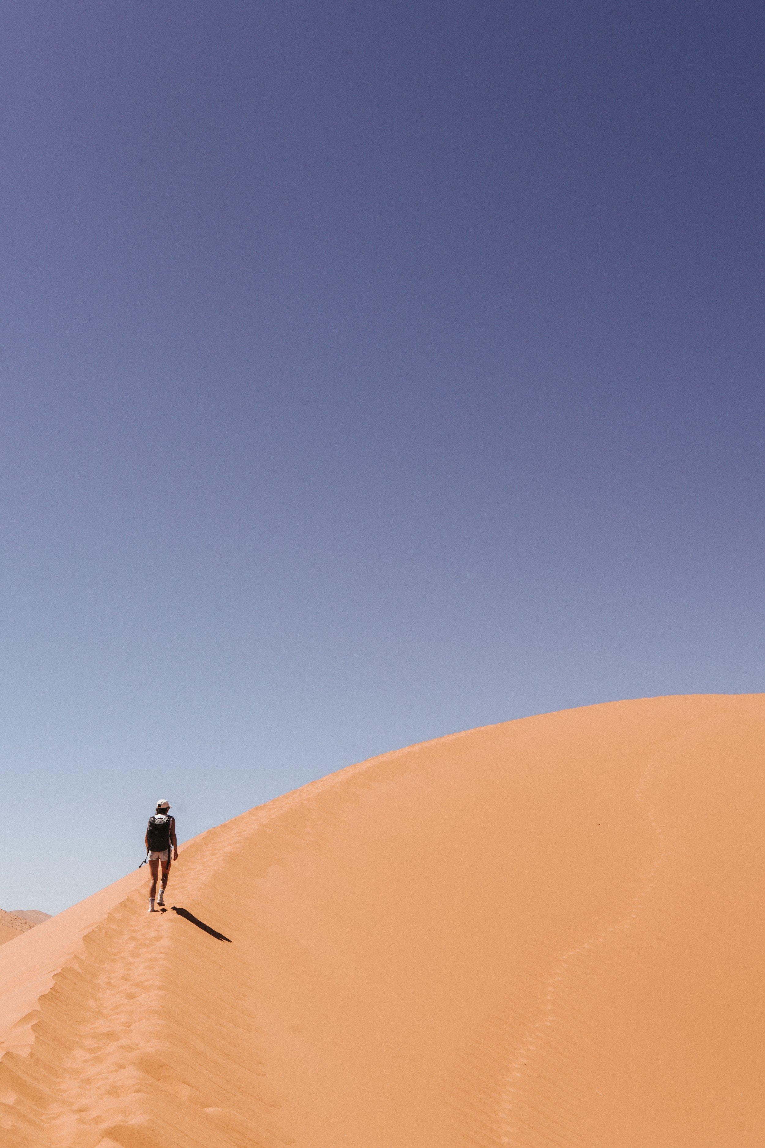 Eine Person in Wanderausrüstung, die einen Sanddünen in einer Wüste entlanggeht, unter einem klaren blauen Himmel.