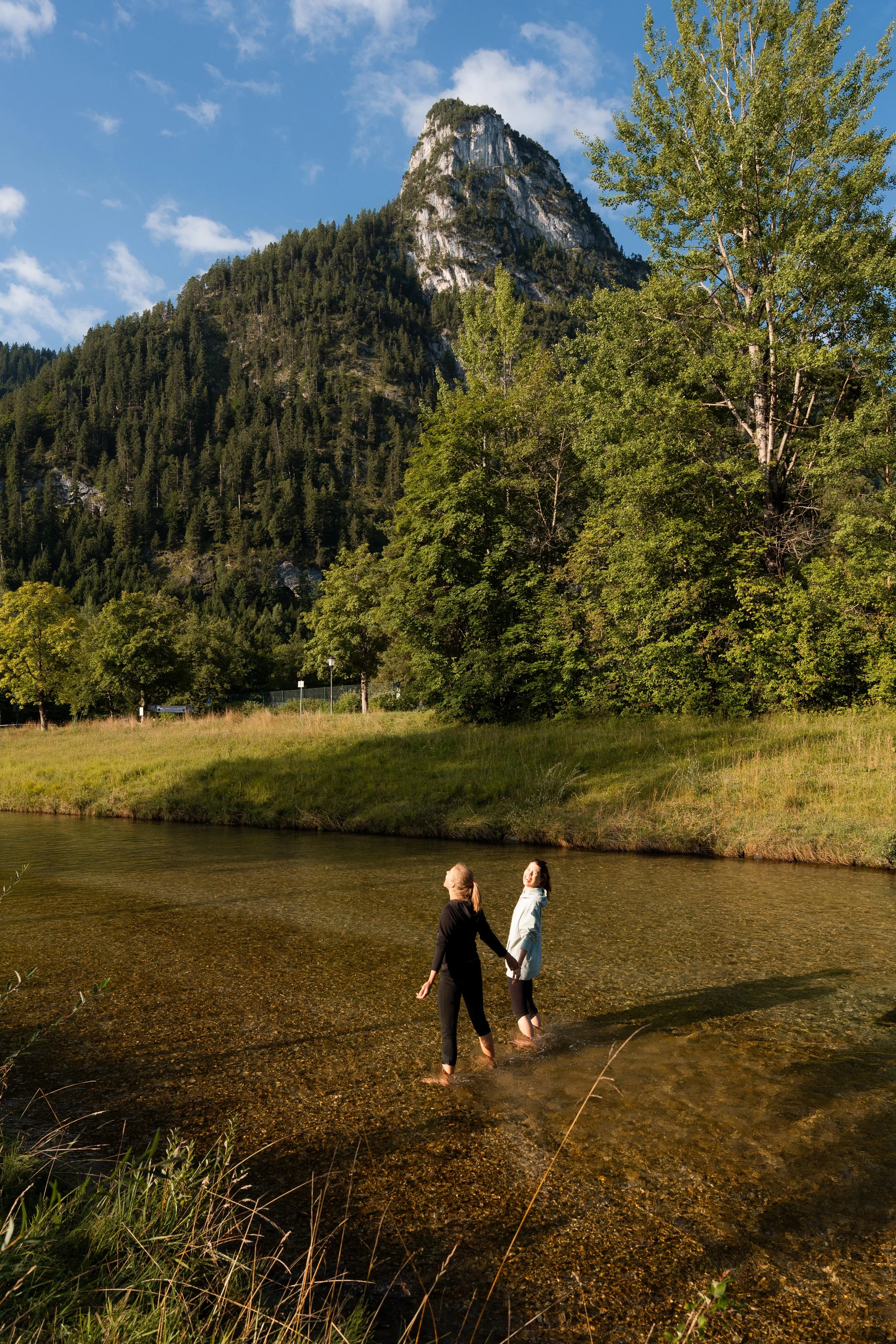 Zwei Frauen stehen barfuß im Fluss, halten sich an den Händen und unterhalten sich mit einem Berg und Bäumen im Hintergrund bei sonnigem Wetter.