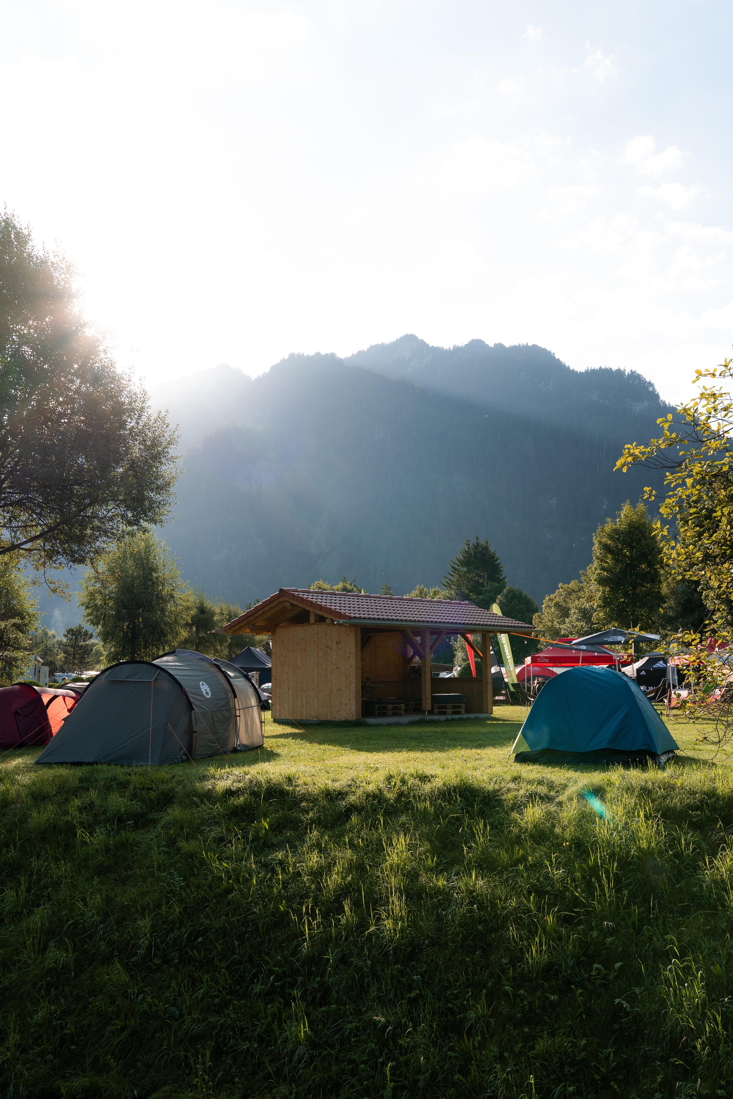 Campingplatz im Grünen mit Zelten, einem kleinen Holzhaus und Blick auf Berge im Hintergrund bei Sonnenschein.