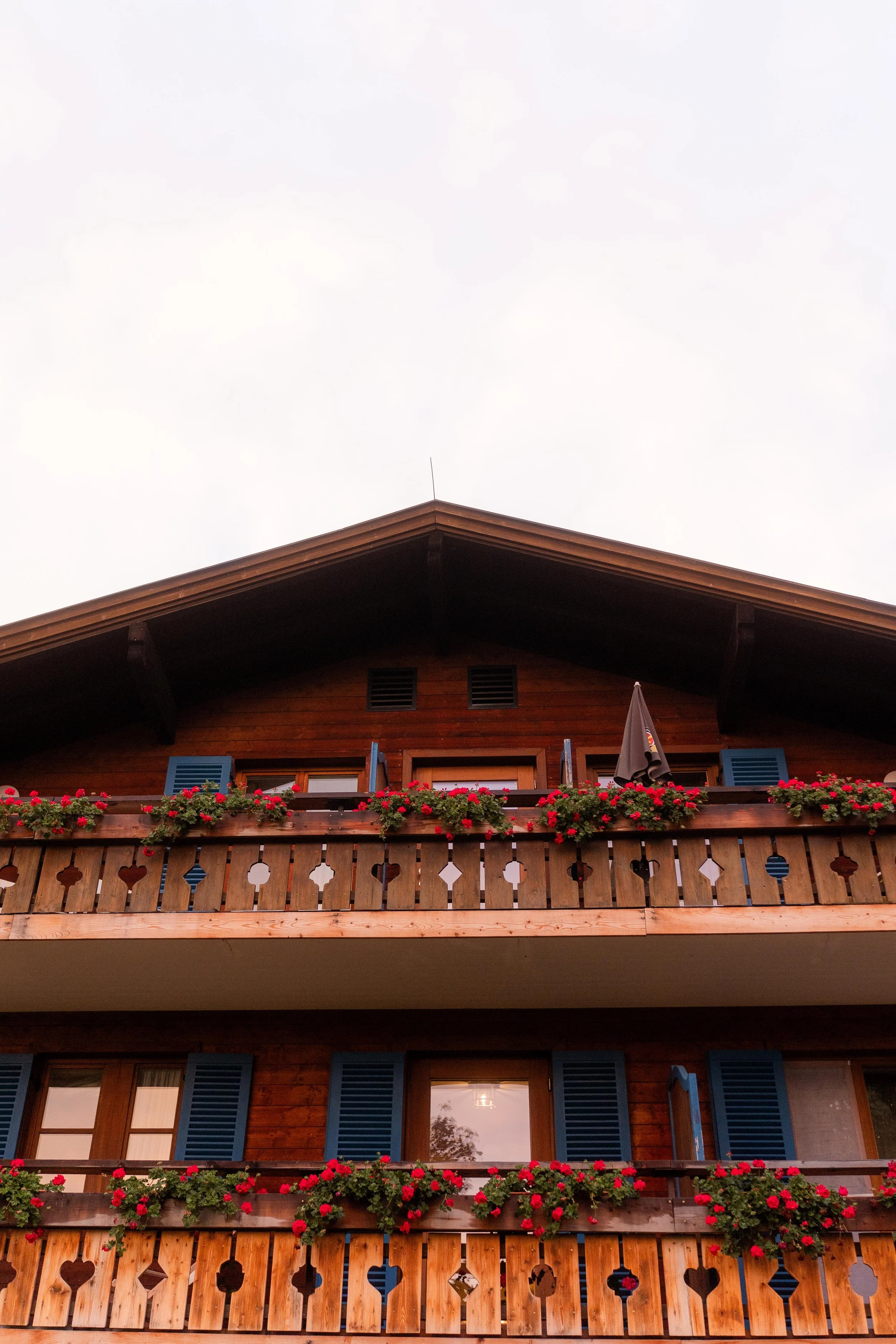 Drei Stockwerke eines Holzhauses mit blauen Fensterläden und roten Blumen auf den Blumenkästen auf den Balkonen.