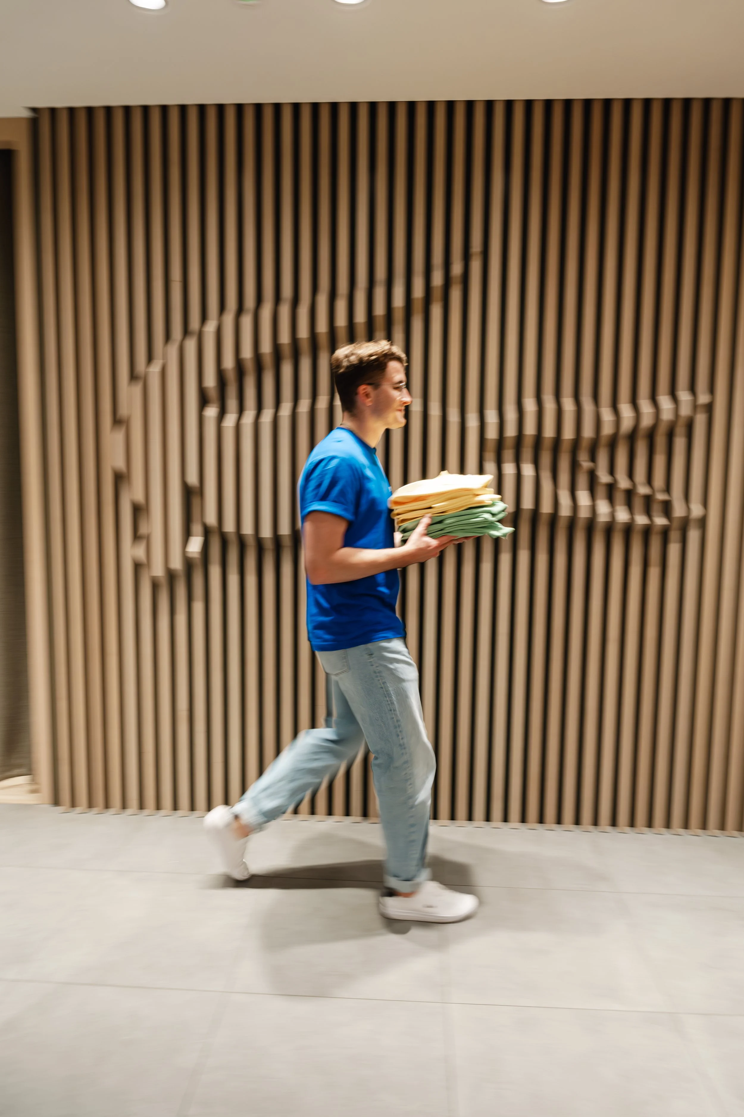 Junge Person in blauer T-Shirt und Jeans trägt drei Stapel Kleidung in einem modernen Raum mit Holzdekoration an der Wand.