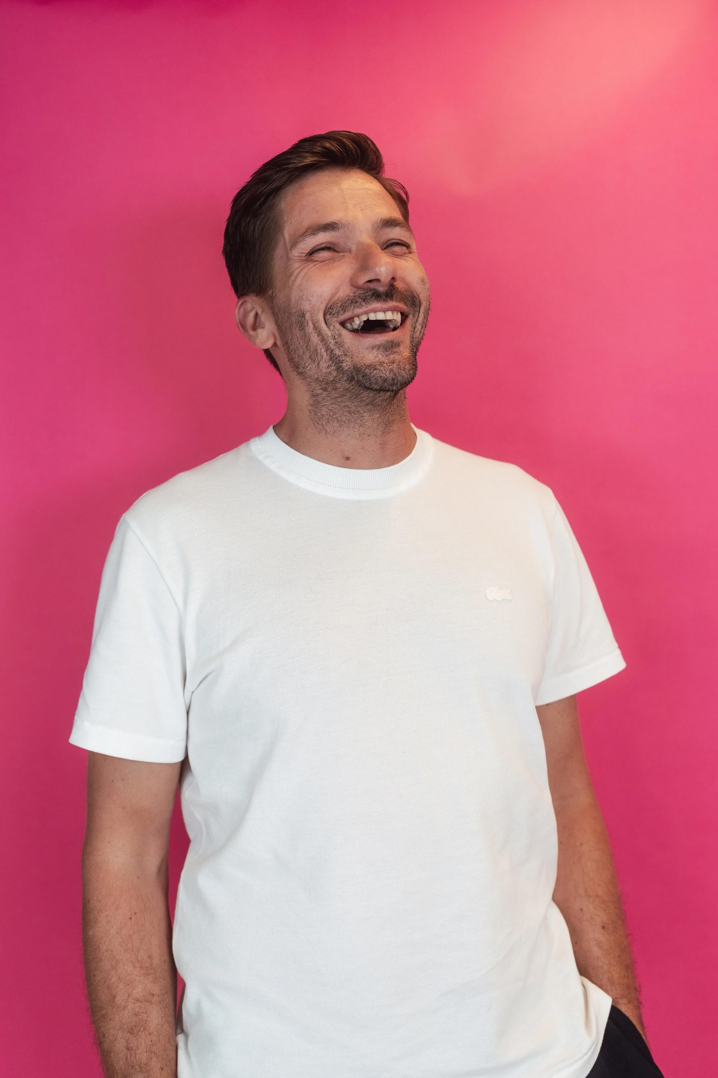 Lächelnder Mann steht vor pinkem Hintergrund, trägt weißen T-Shirt.