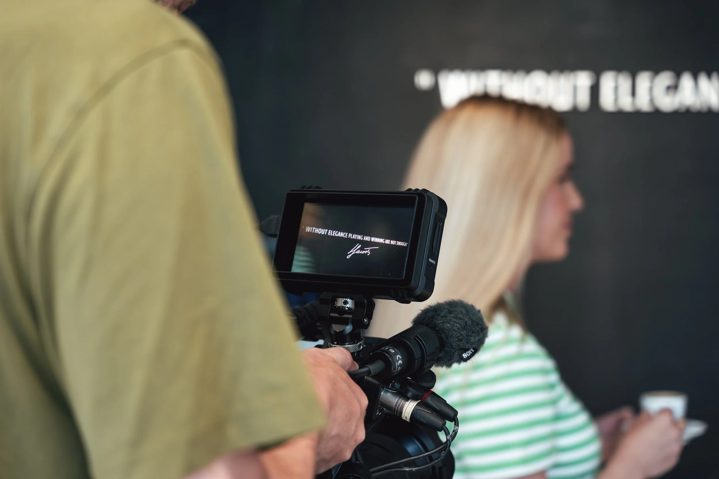 Ein Mann filmt eine blonde Frau mit einem Videokamera, im Hintergrund ist schwarzer Wand mit weißen Text.