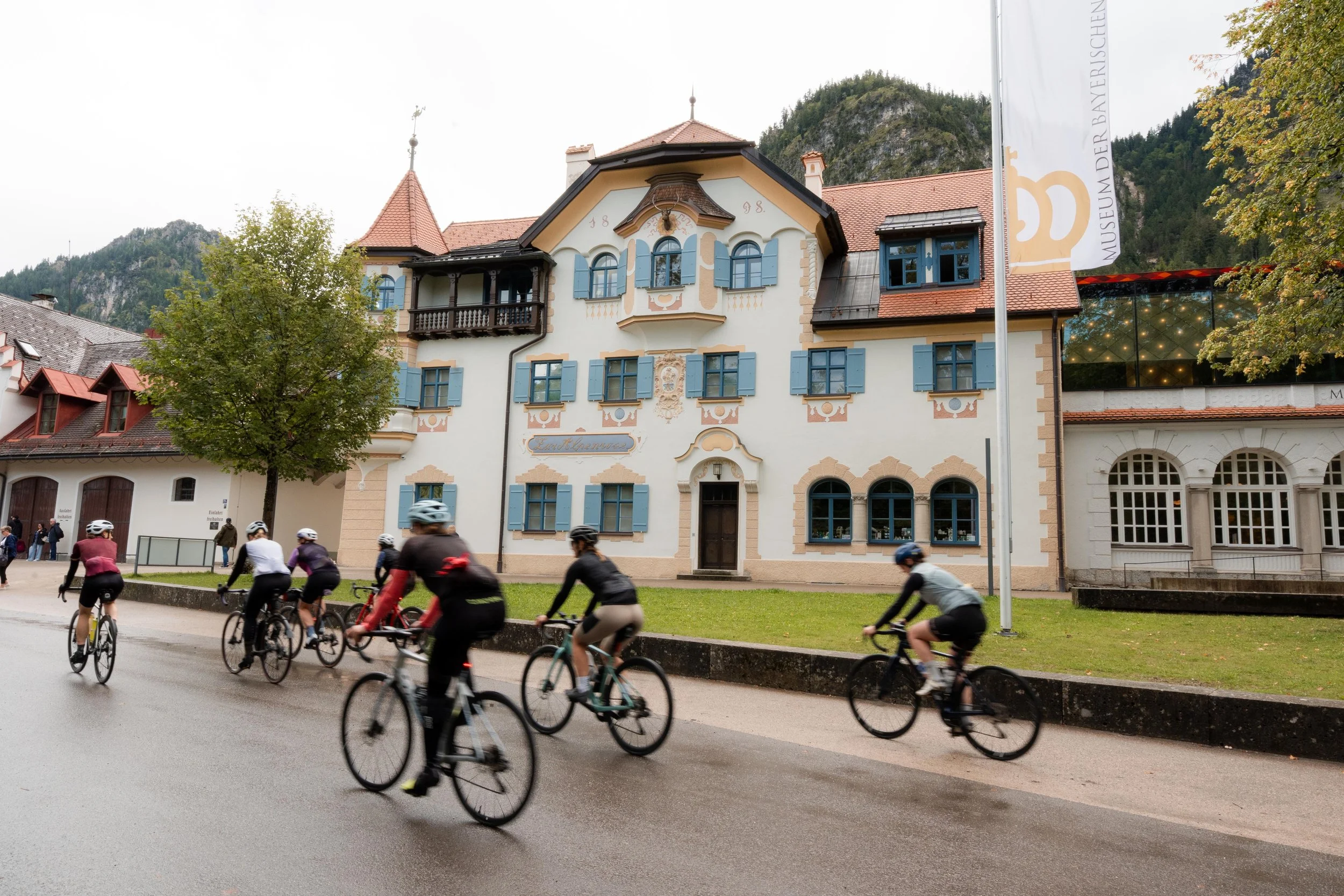 Eine Gruppe von Radfahrern fährt an einem historischen Gebäude mit blau gestrichenen Fensterläden vorbei, im Hintergrund Berge, an einem bewölkten Tag.