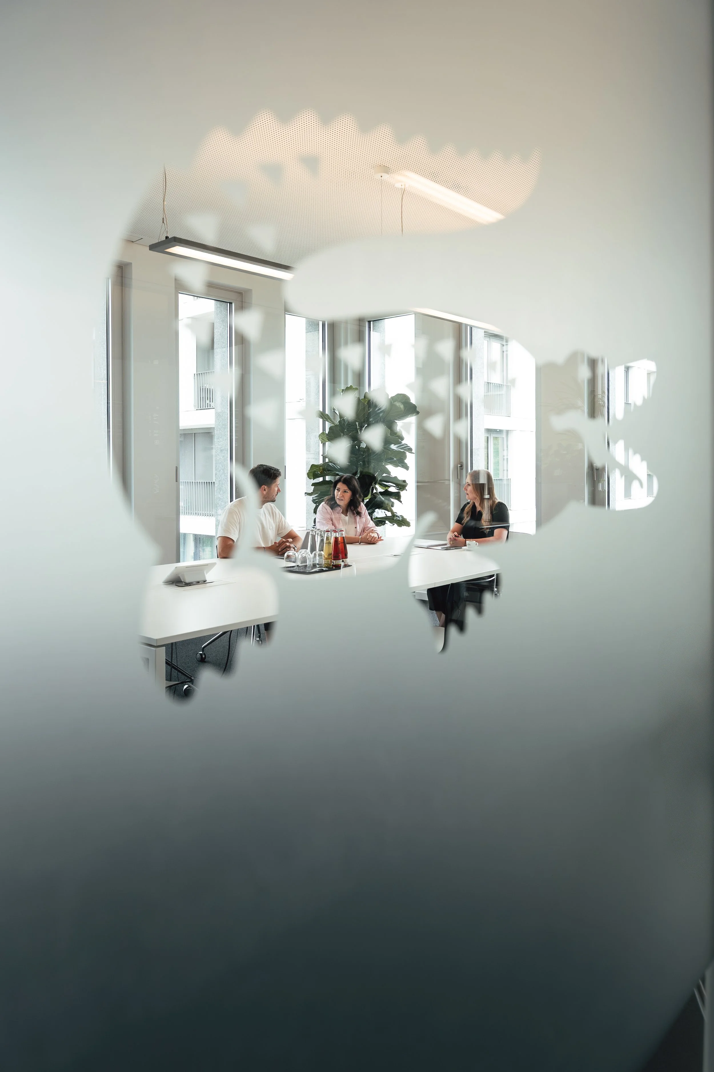 Drei Personen sitzen im Besprechungsraum an einem weißen Tisch, beobachtet durch eine Aussparung in einer Tür oder Wand, mit großen Fenstern im Hintergrund und einer großen Pflanze in der Ecke.