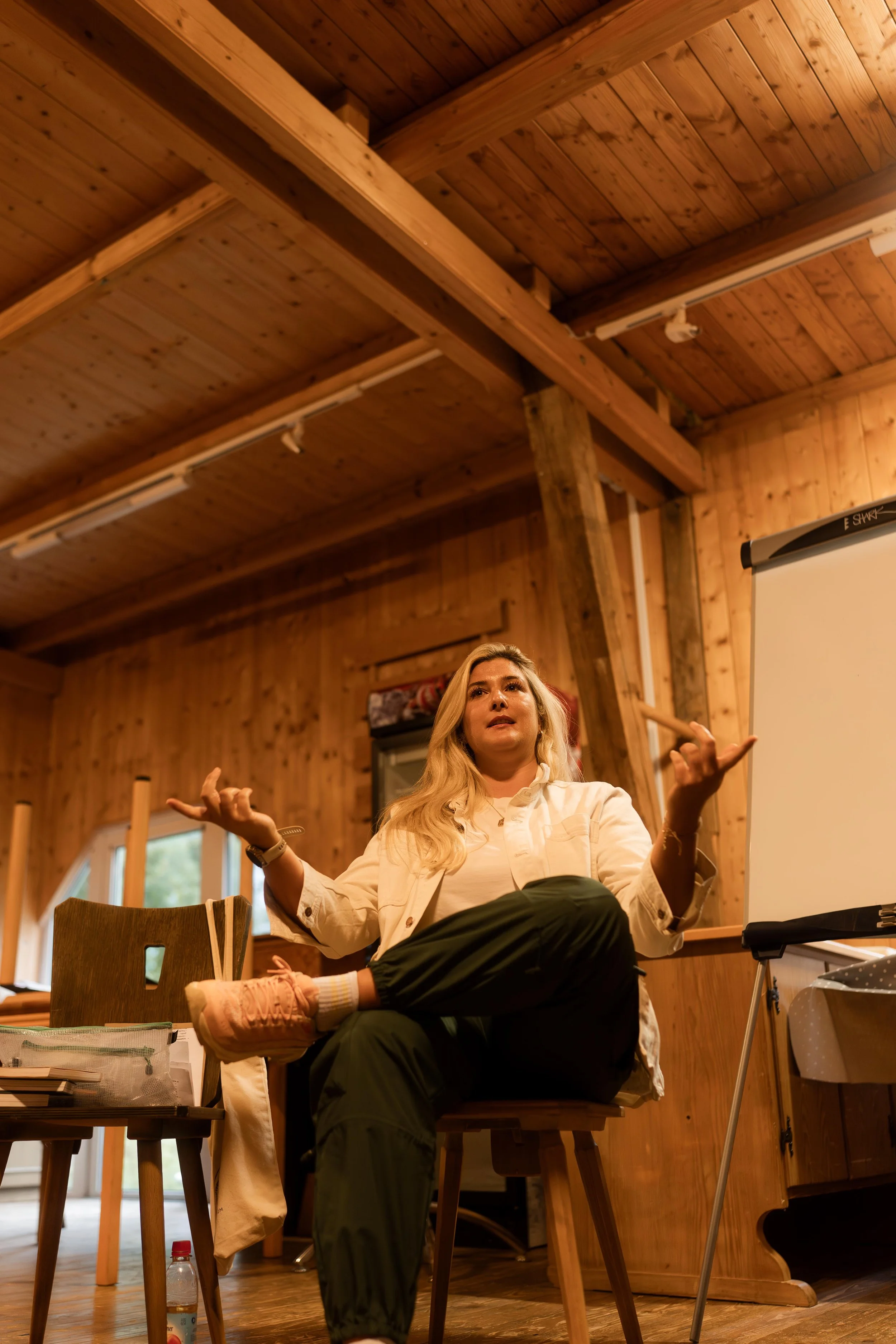 Eine Frau sitzt in einem Raum mit Holzwänden und -decke, spricht und gestikuliert während einer Präsentation oder Diskussion, neben einem Flipchart.