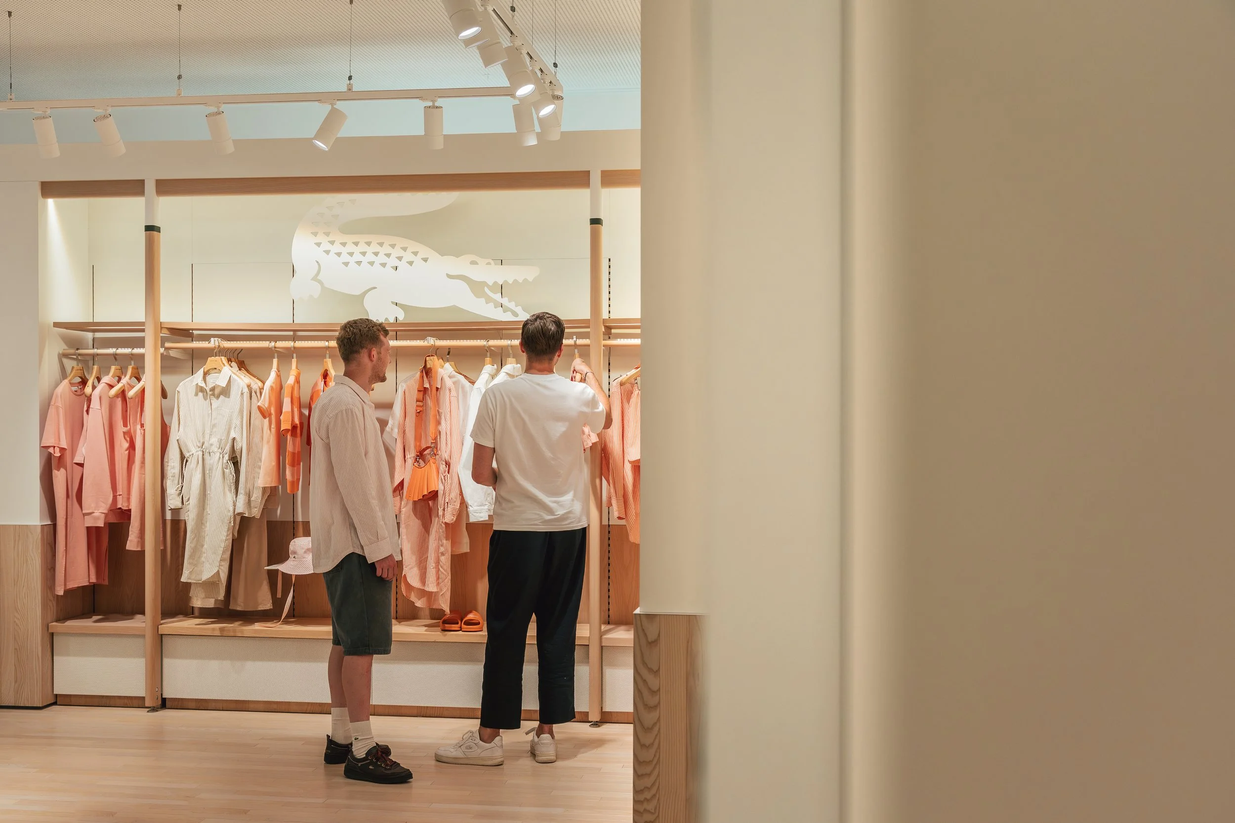 Zwei Männer schauen sich in einem Kleidungsgeschäft an, das hauptsächlich hellfarbige Kleidung in Pastelltönen verkauft, mit einem Krokodil-Logo im Hintergrund.