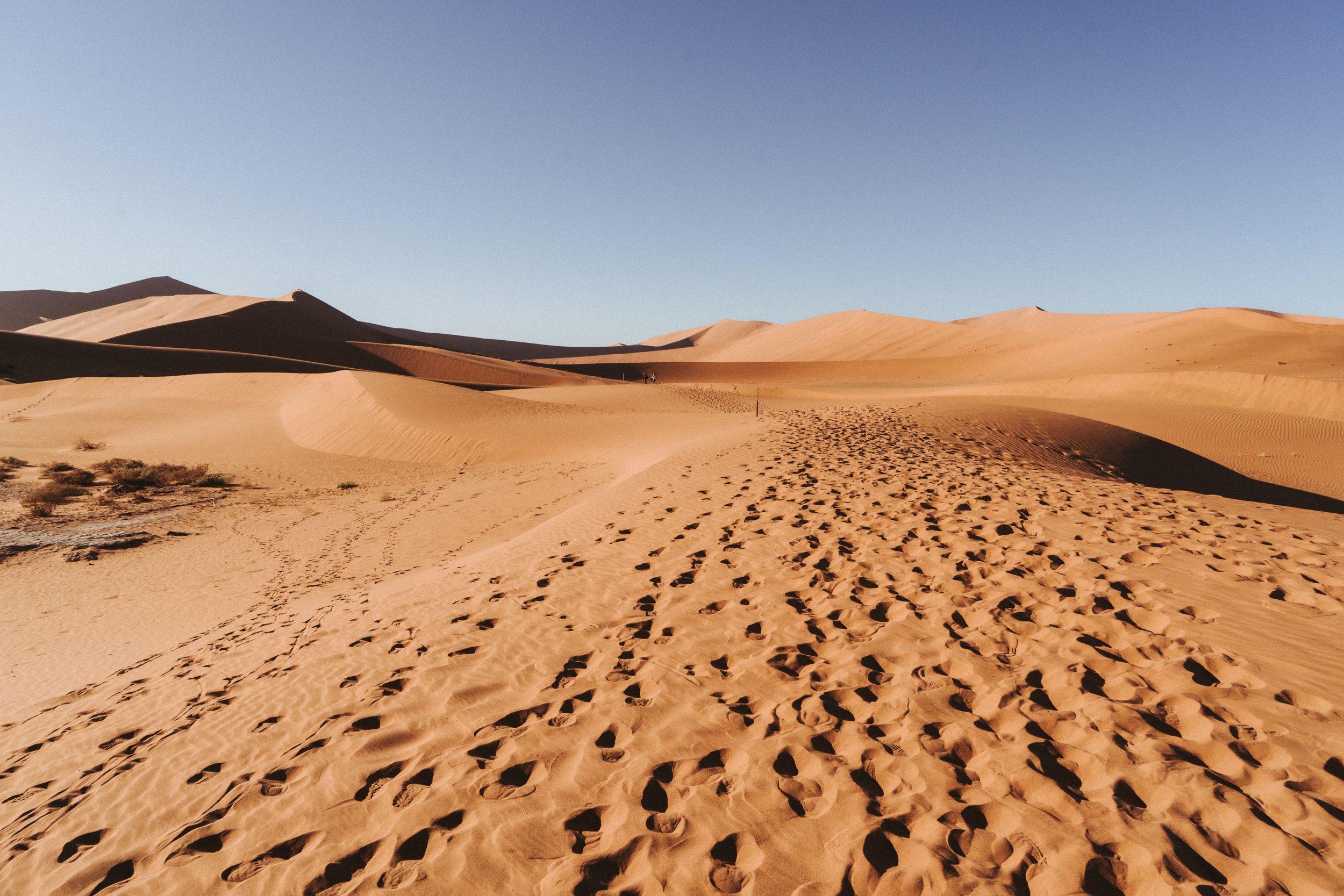 Wüstenlandschaft mit Sanddünen und Fußspuren im Sand, unter klarem blauen Himmel.