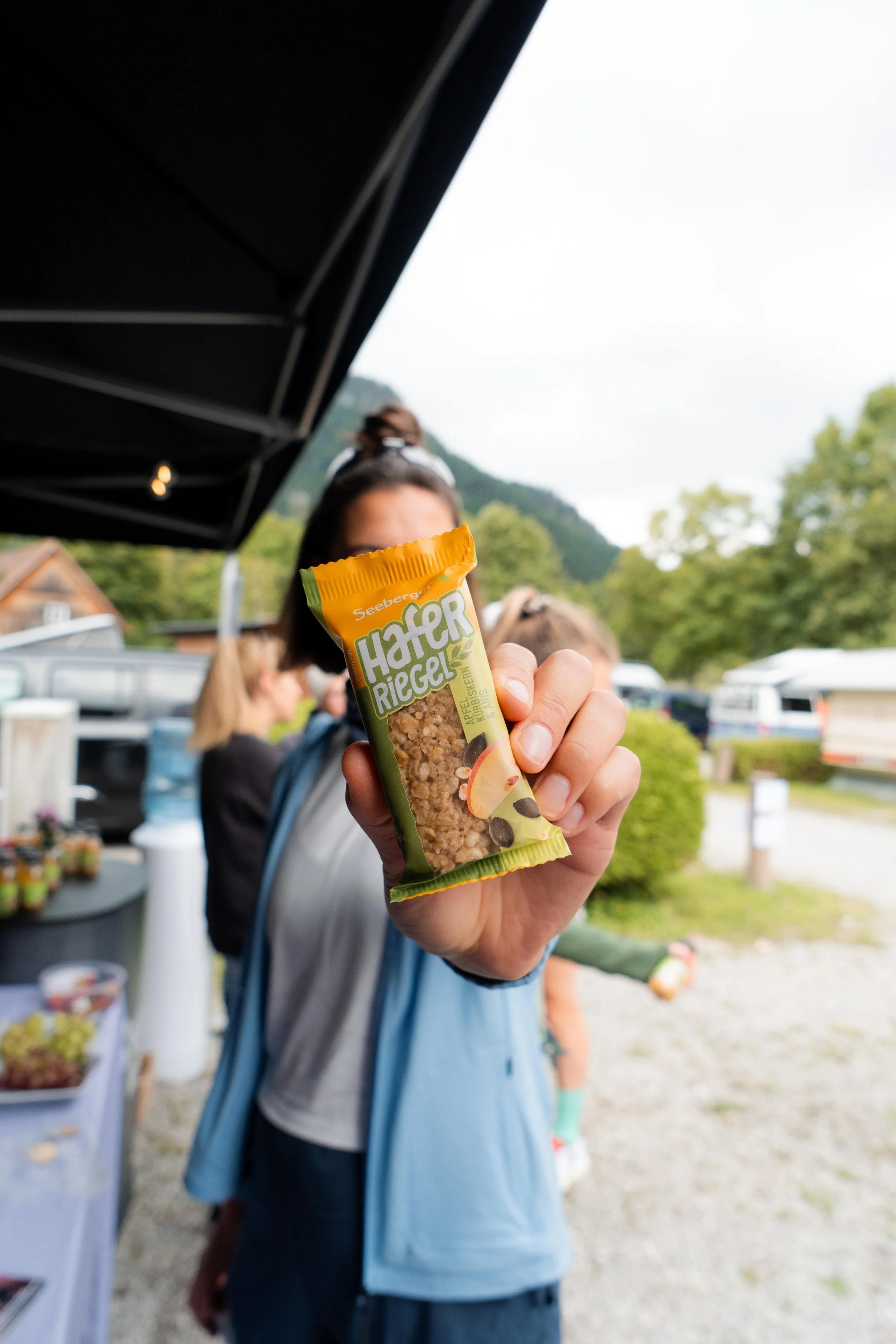 Person hält eine Packung Haferriegel-Knusperriegel in die Kamera, im Hintergrund sind weitere Menschen und Natur zu sehen.