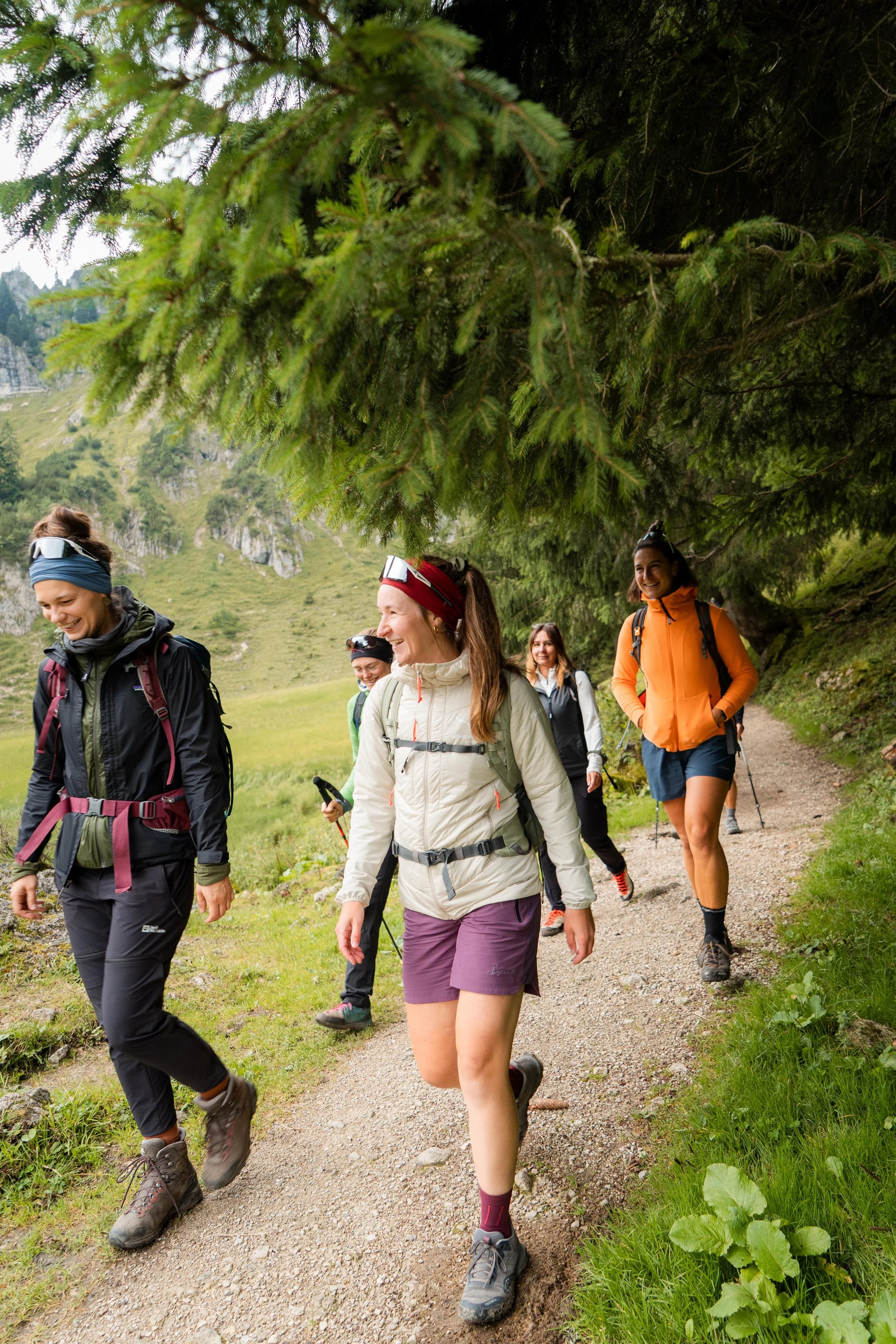 Eine Gruppe von fünf Frauen beim Wandern auf einem schmalen Waldweg in einer grünen, bergigen Landschaft, mit Bäumen und Felsen im Hintergrund.
