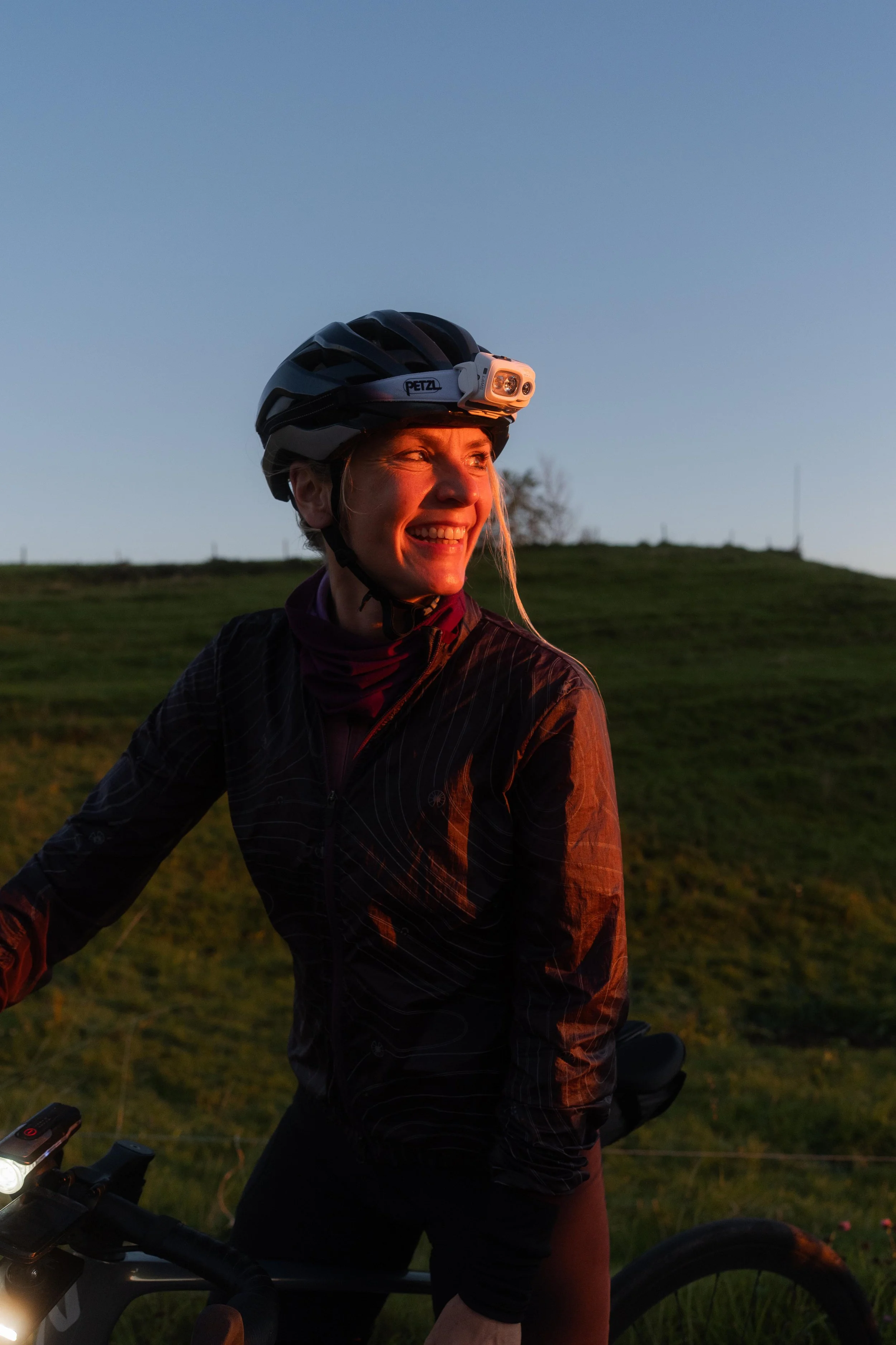 Lächelnde Frau mit Fahrradhelm und Stirnlampe auf Mountainbike in ländlicher Landschaft bei Sonnenuntergang.