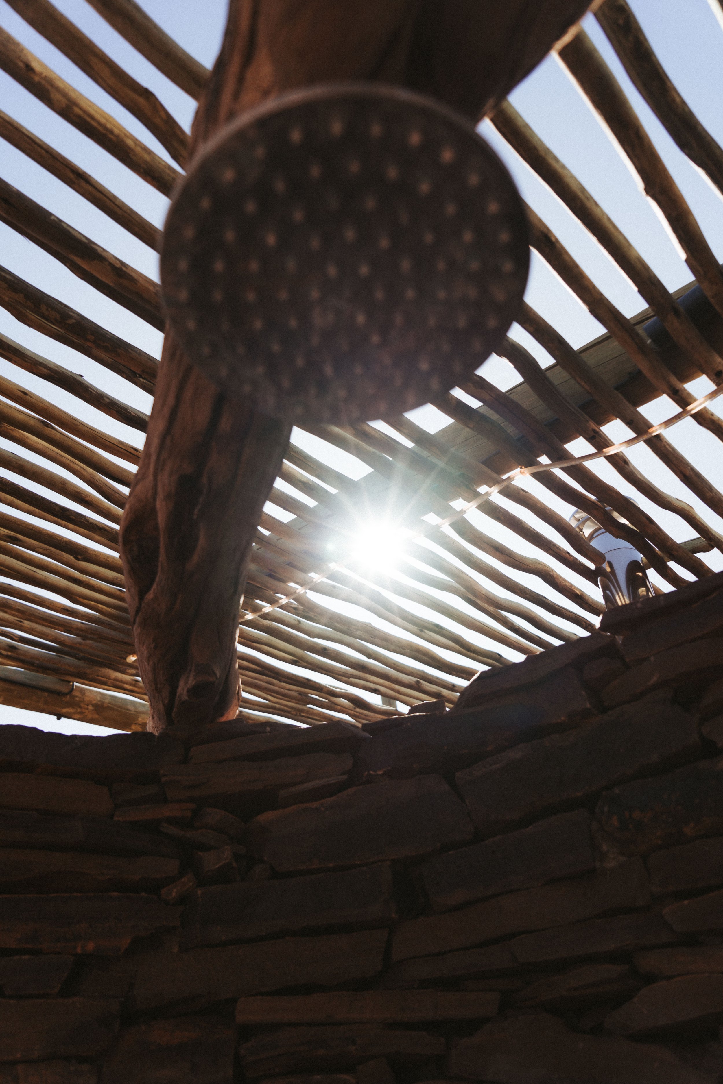 Blick nach oben auf eine Holzdecke mit Sonnenlicht, geschoßen auf eine Holzsäule, mit Steinen ausgefüllte Wand im Vordergrund, und eine Laterne im Hintergrund.