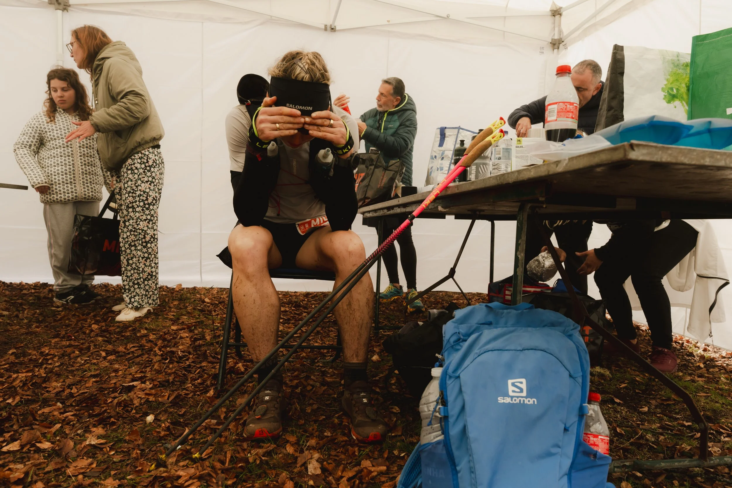 Innenansicht eines Zeltes mit mehreren Menschen, einer sitzt mit Laufbekleidung und Stirnband, während andere bei Ess- und Trinkvorgängen beschäftigt sind; im Hintergrund liegen Utensilien und Getränke.