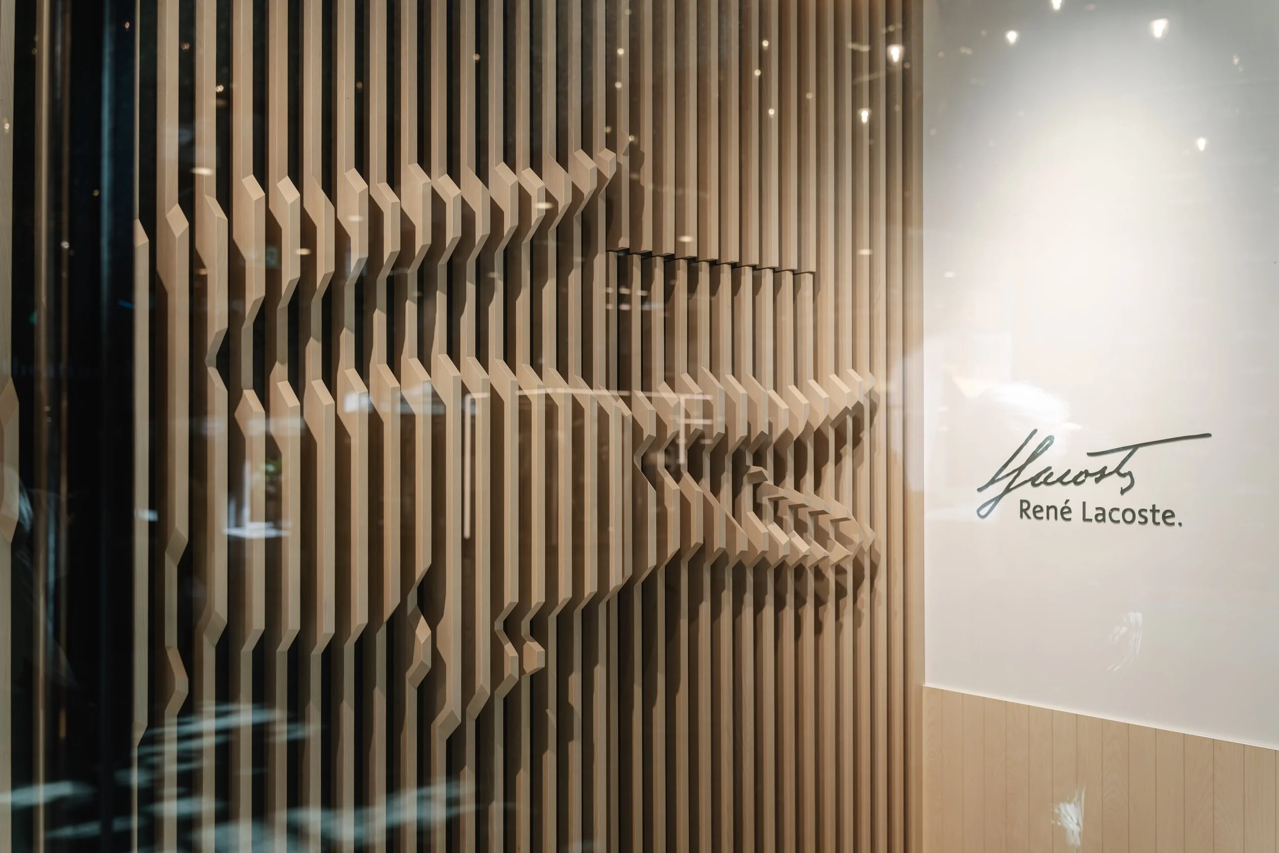 Holzwand mit geschwungenen vertikalen Lamellen und einem Signaturlogo von René Lacoste an einer weißen Wand.