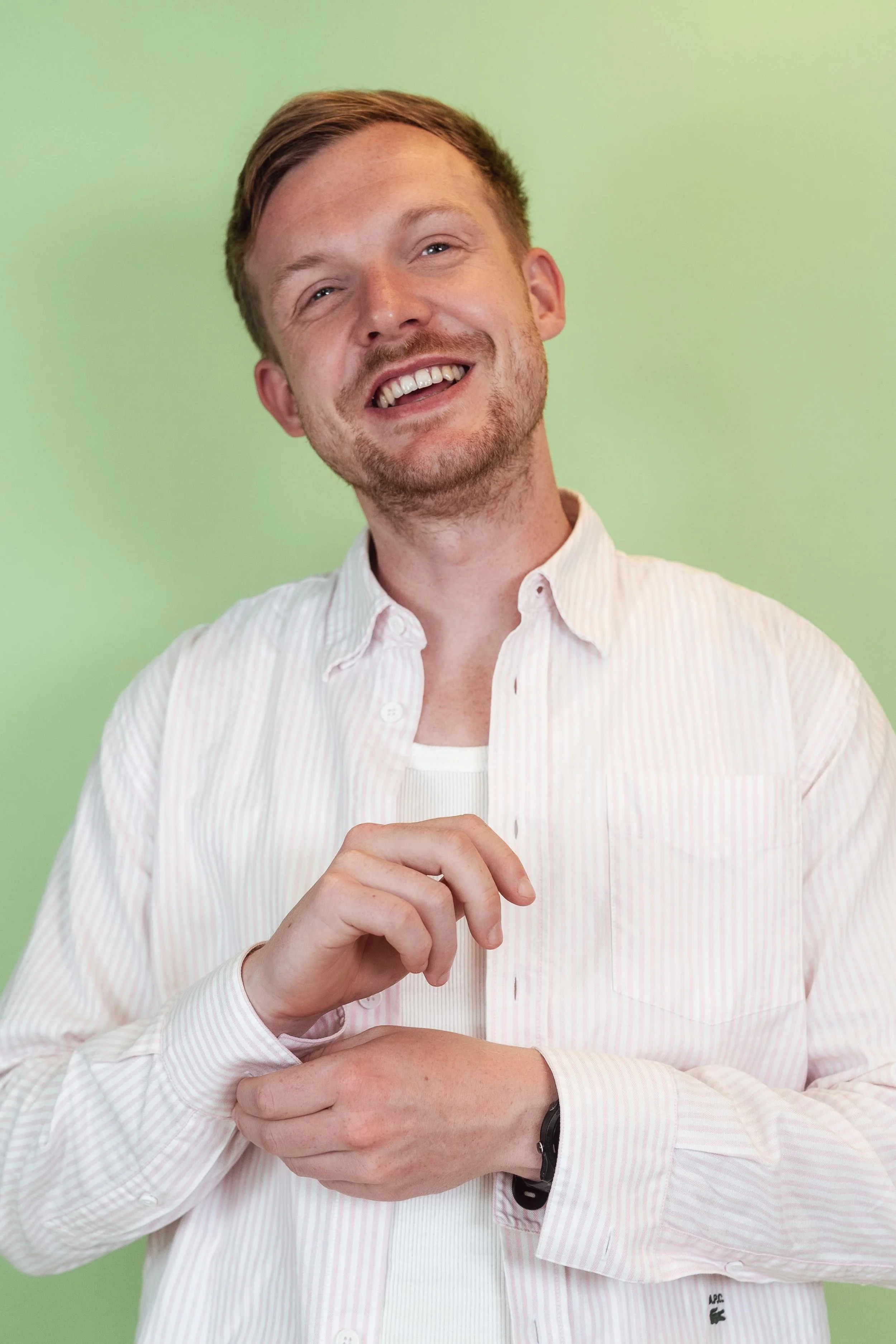 Ein lachender Mann mit hellbraunen Haaren vor einem grünen Hintergrund, trägt ein weißes Hemd, und schaut fröhlich in die Kamera.