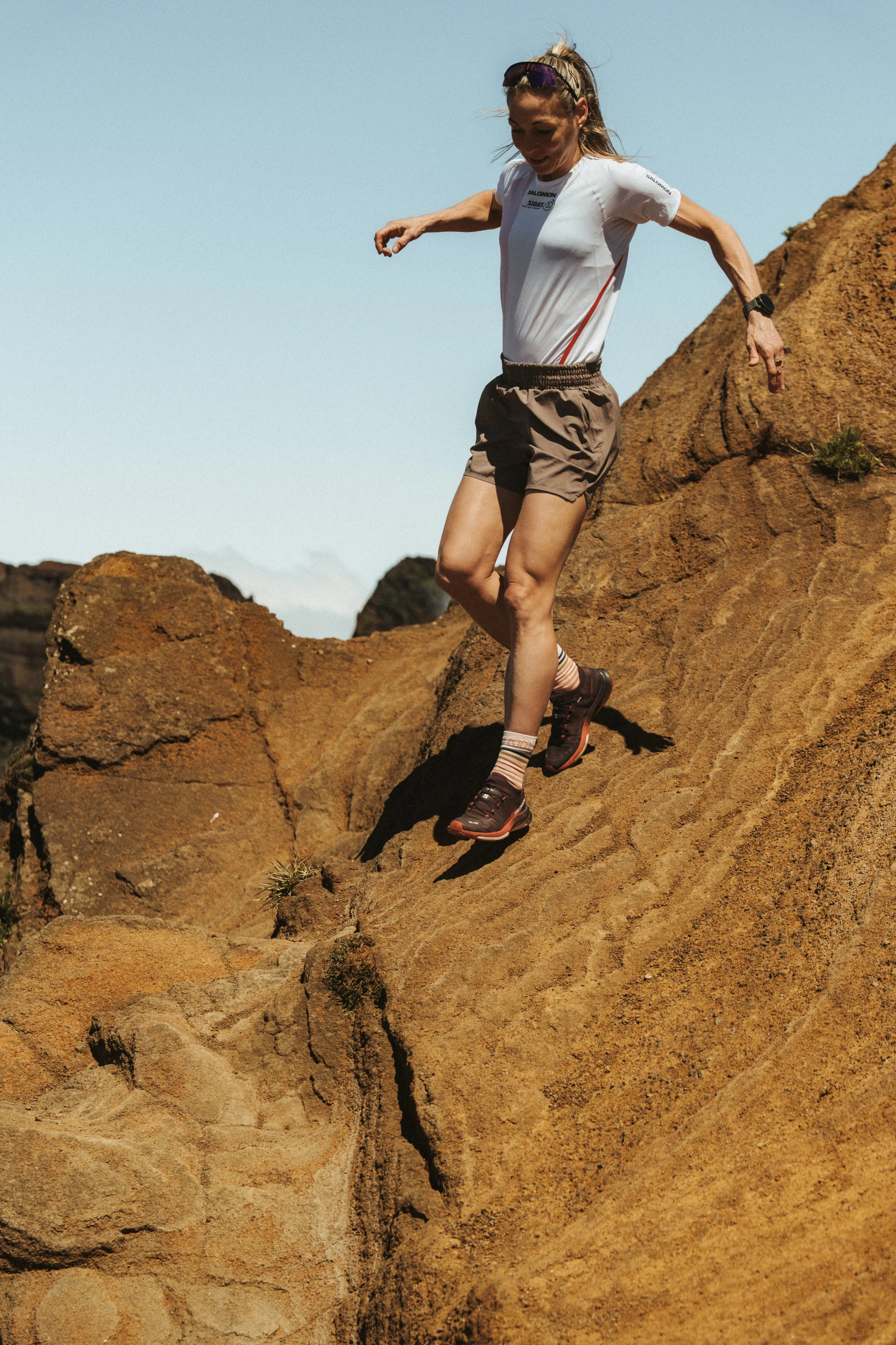 Frau beim Klettern auf Felsen im Freien bei sonnigem Wetter, trägt sportliche Kleidung und Wanderschuhe.