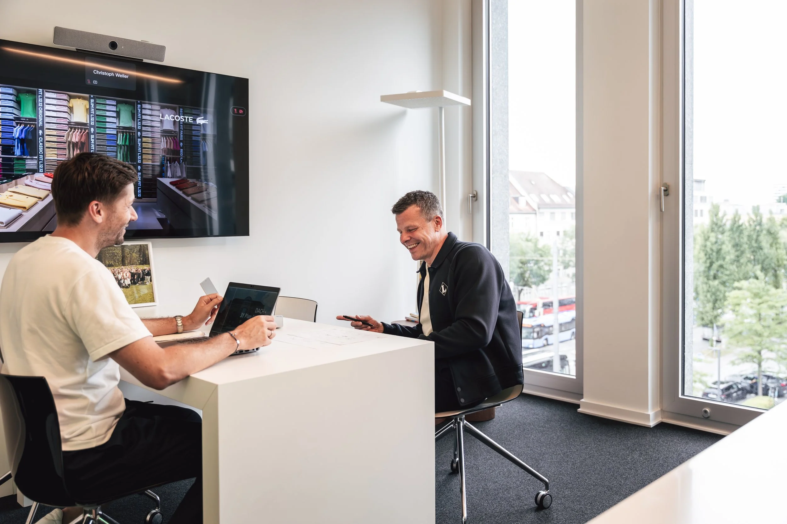 Zwei Männer sitzen an einem weißen Tisch in einem gut beleuchteten Büro, lachen und unterhalten sich, während sie mit Smartphones und einem Laptop arbeiten. Ein großes Fenster bietet einen Blick auf eine Stadt.