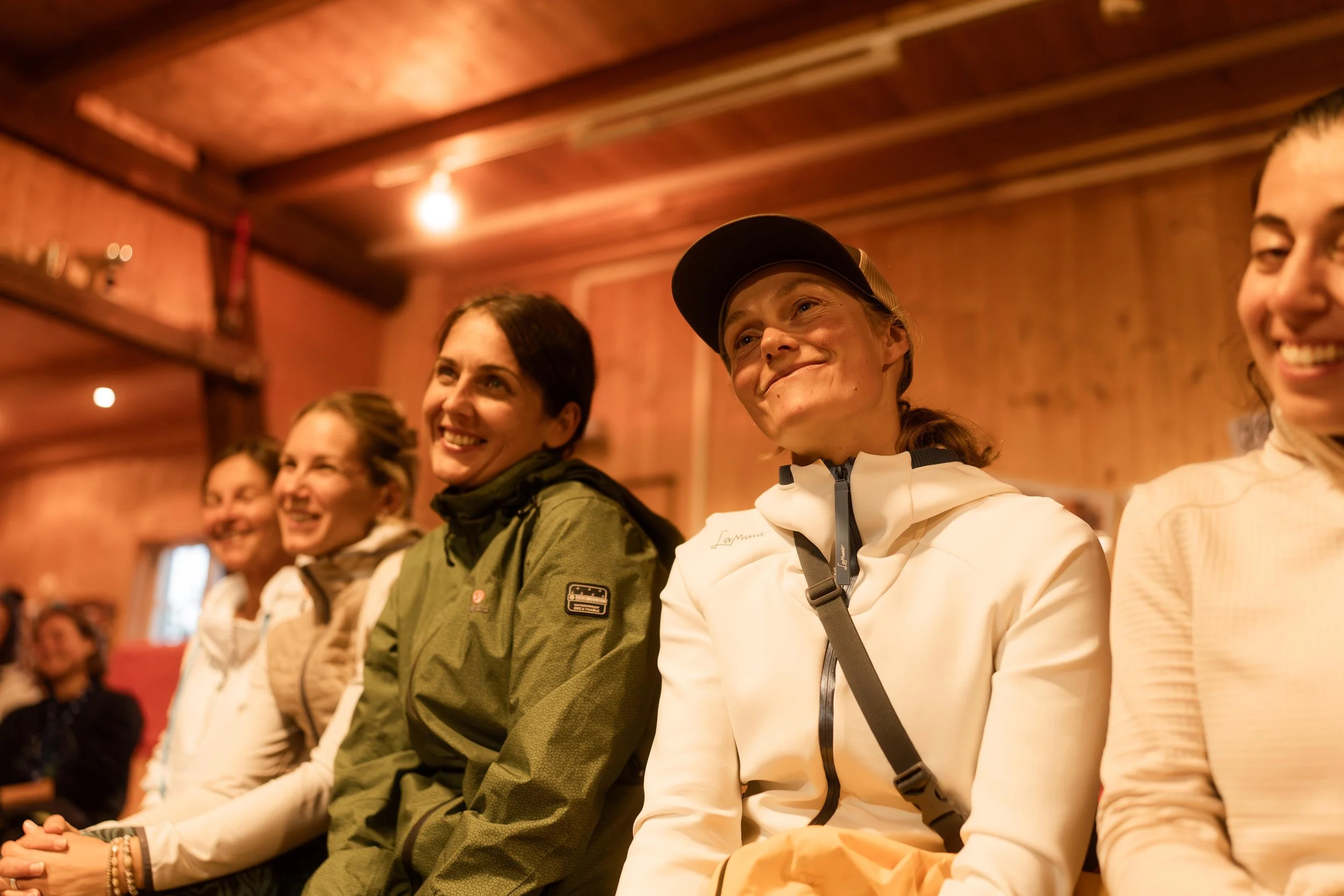 Eine Gruppe von Frauen, die in einem rustikalen Holzraum sitzen und lachen, während sie einem Vortrag oder einer Präsentation zuhören.