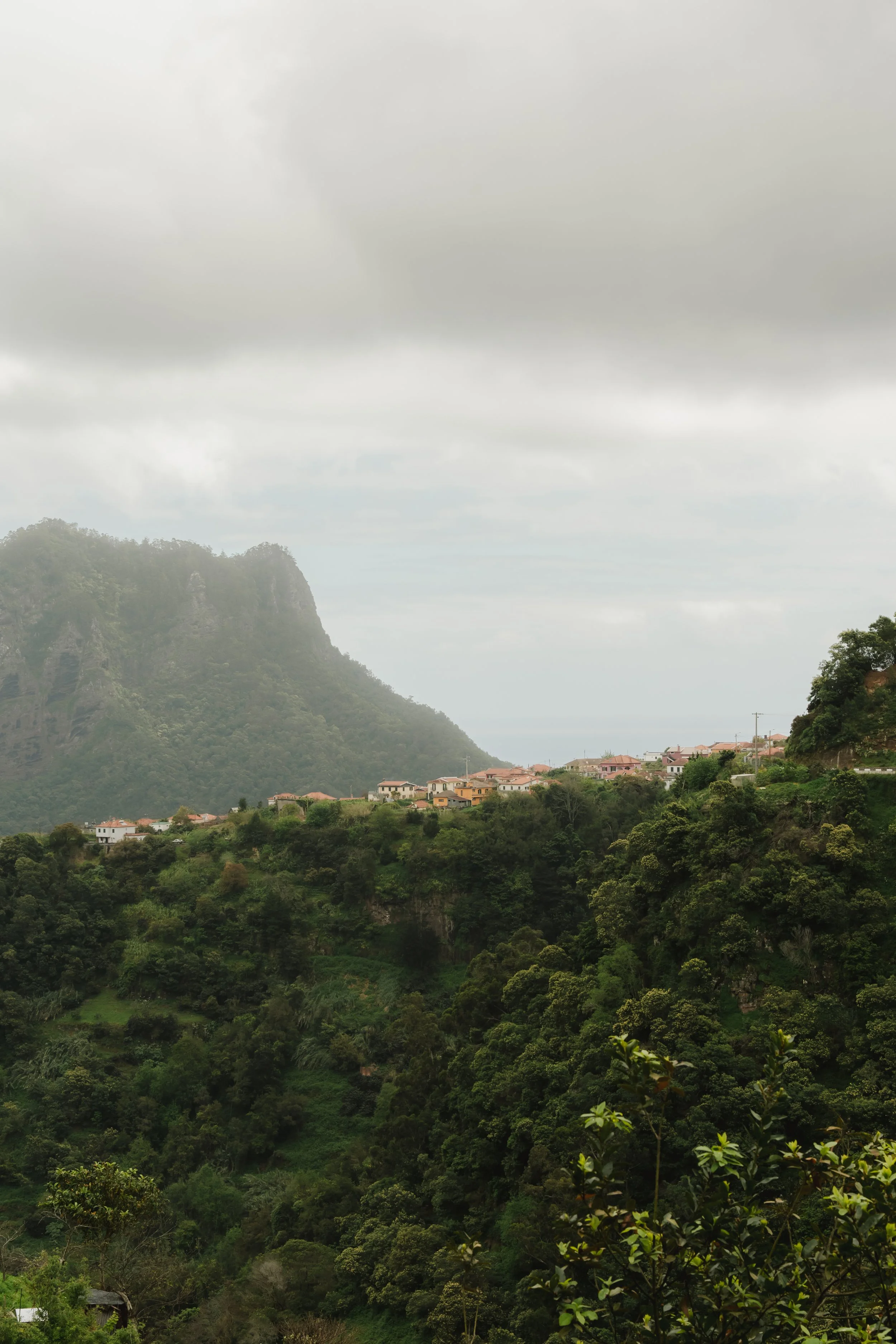 Eine Landschaft mit bewaldeten Hügeln, einem Dorf auf der Hügelkuppe und einer Bergkette im Hintergrund bei bewölktem Himmel.