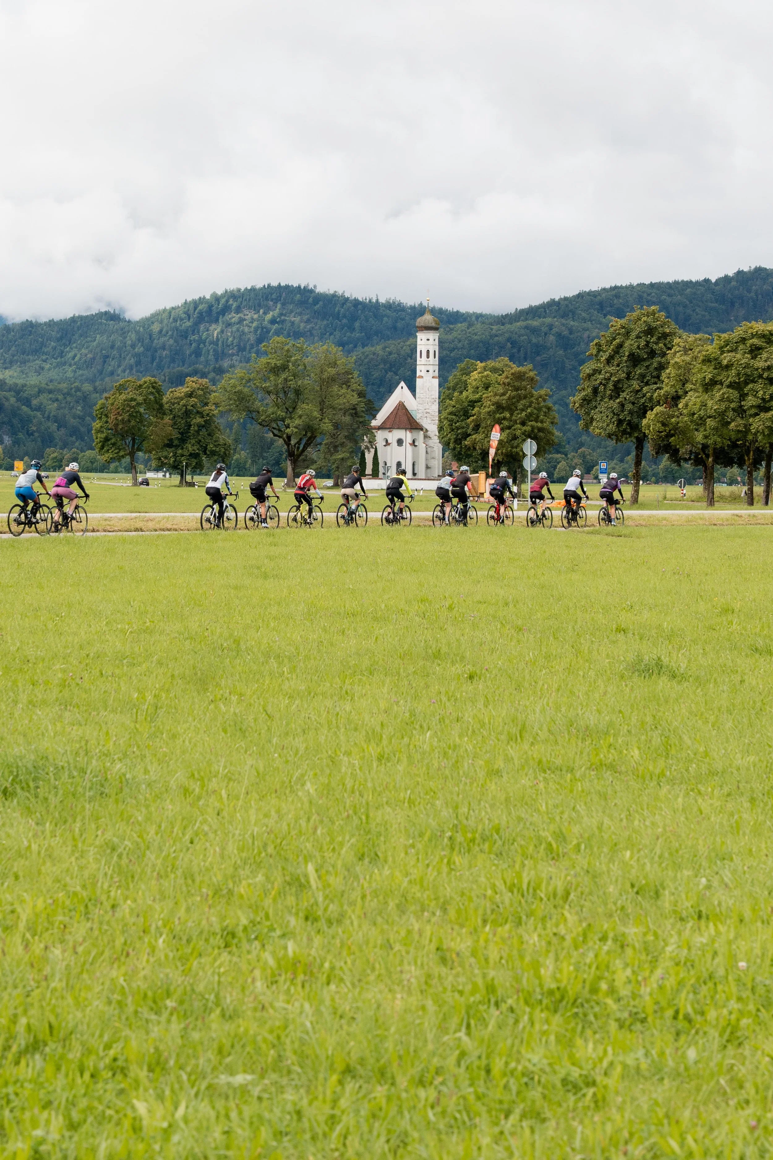 Eine Gruppe von Radfahrern fährt durch eine grüne Wiese mit Bäumen und einer kleinen weißen Kirche im Hintergrund, vor Berglandschaft und bewölktem Himmel.