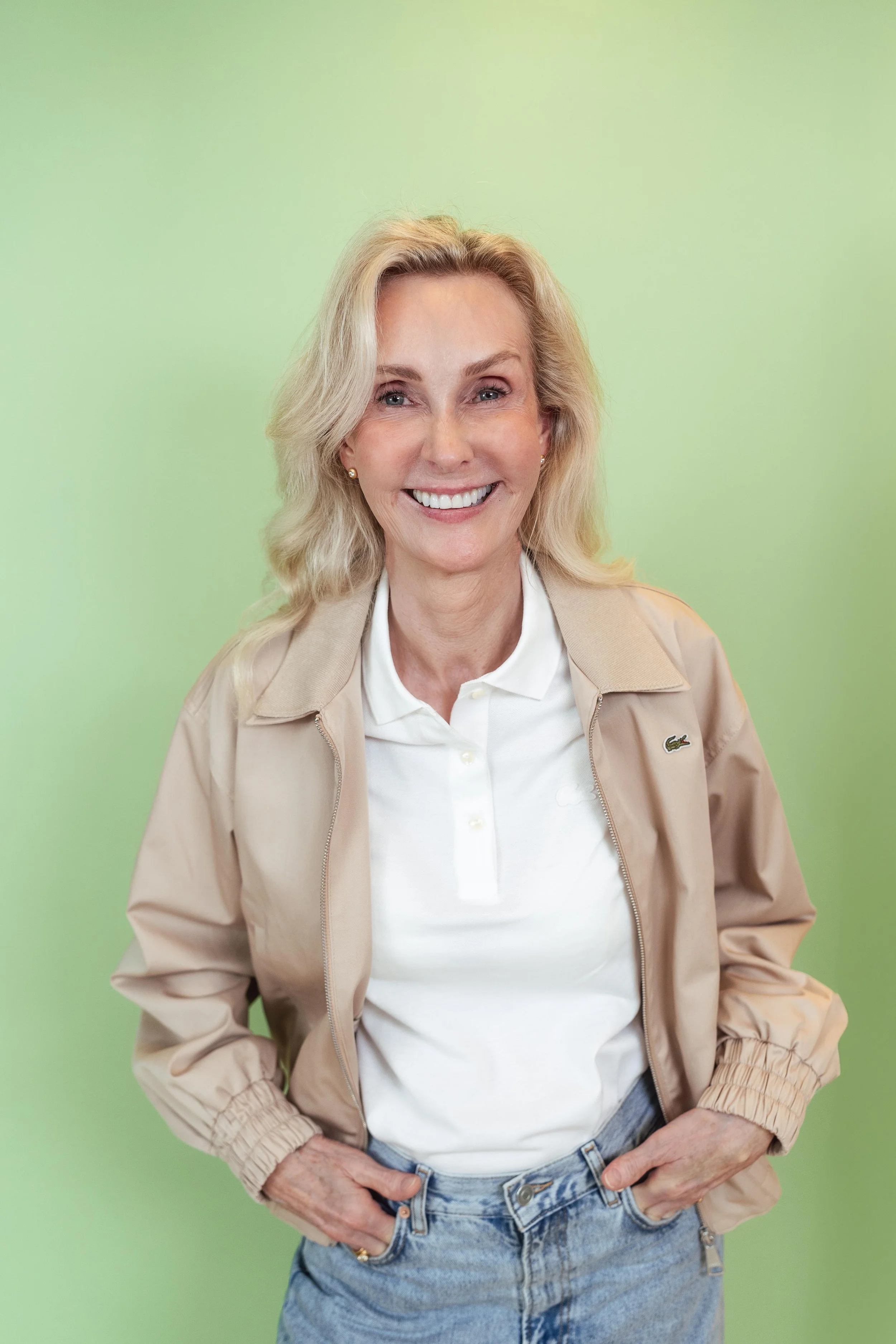 Eine lächelnde Frau mit blonden Haaren in einem beige Jacke und weißen Hemd vor einem hellgrünen Hintergrund.