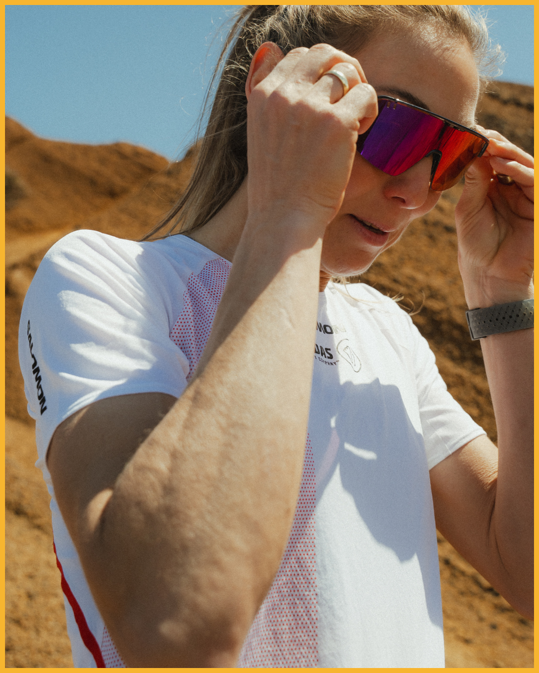 Eine Frau beim Sport trägt rote Sonnenbrille, weißen Sport-Top, graue Uhr, in einer Wüstenlandschaft bei sonnigem Wetter.